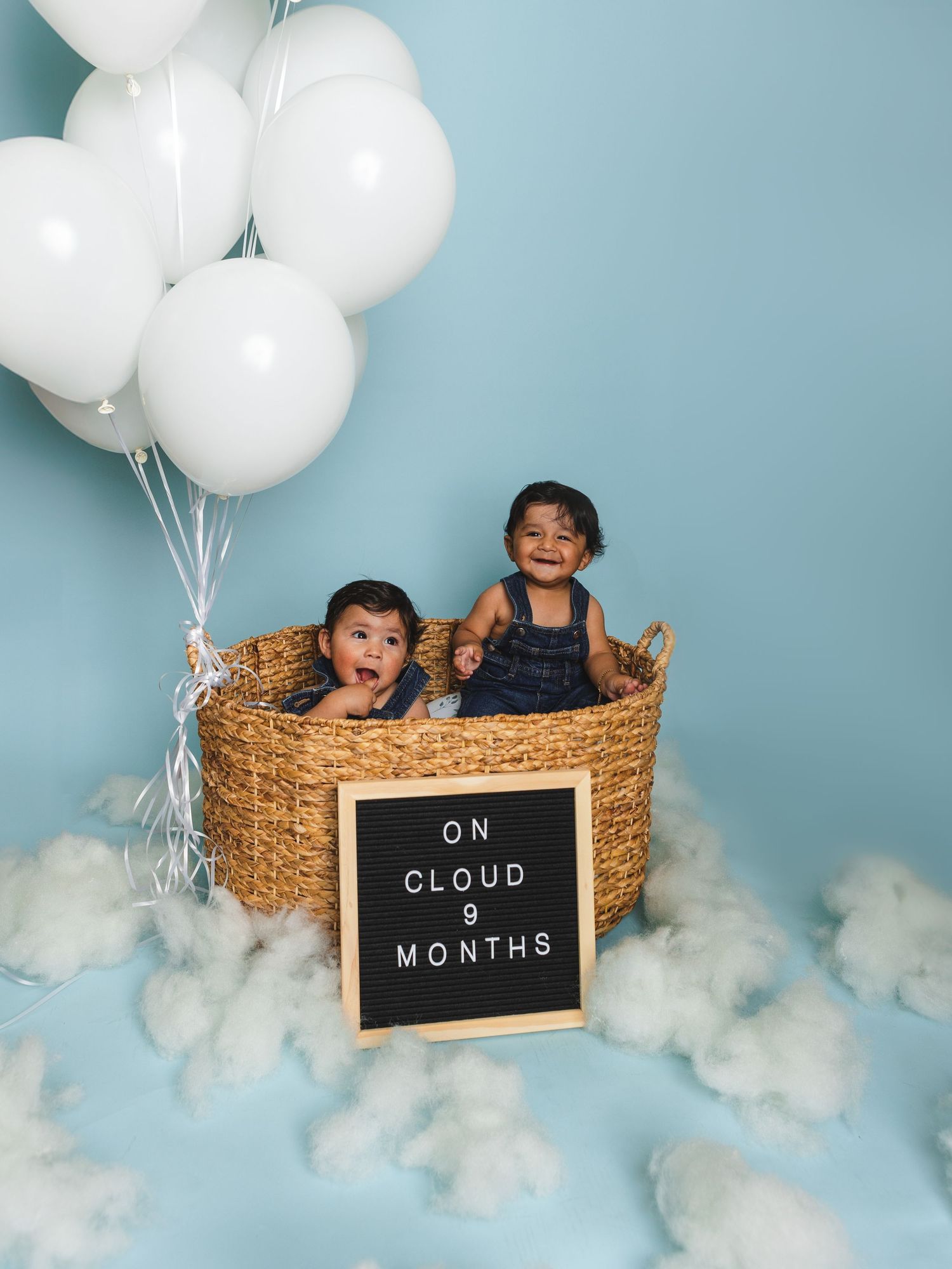 Playful baby photo session featuring white balloons and a rustic basket with cloud decorations.