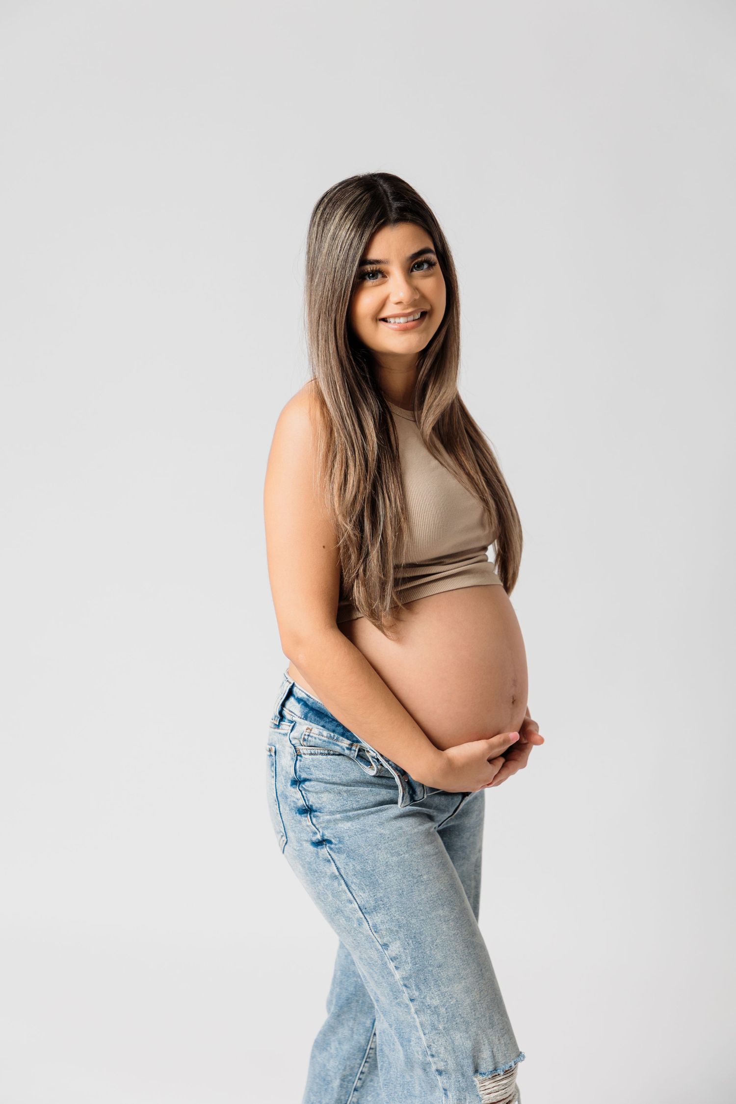 A maternity photoshoot featuring casual denim and a beige top against a white backdrop.