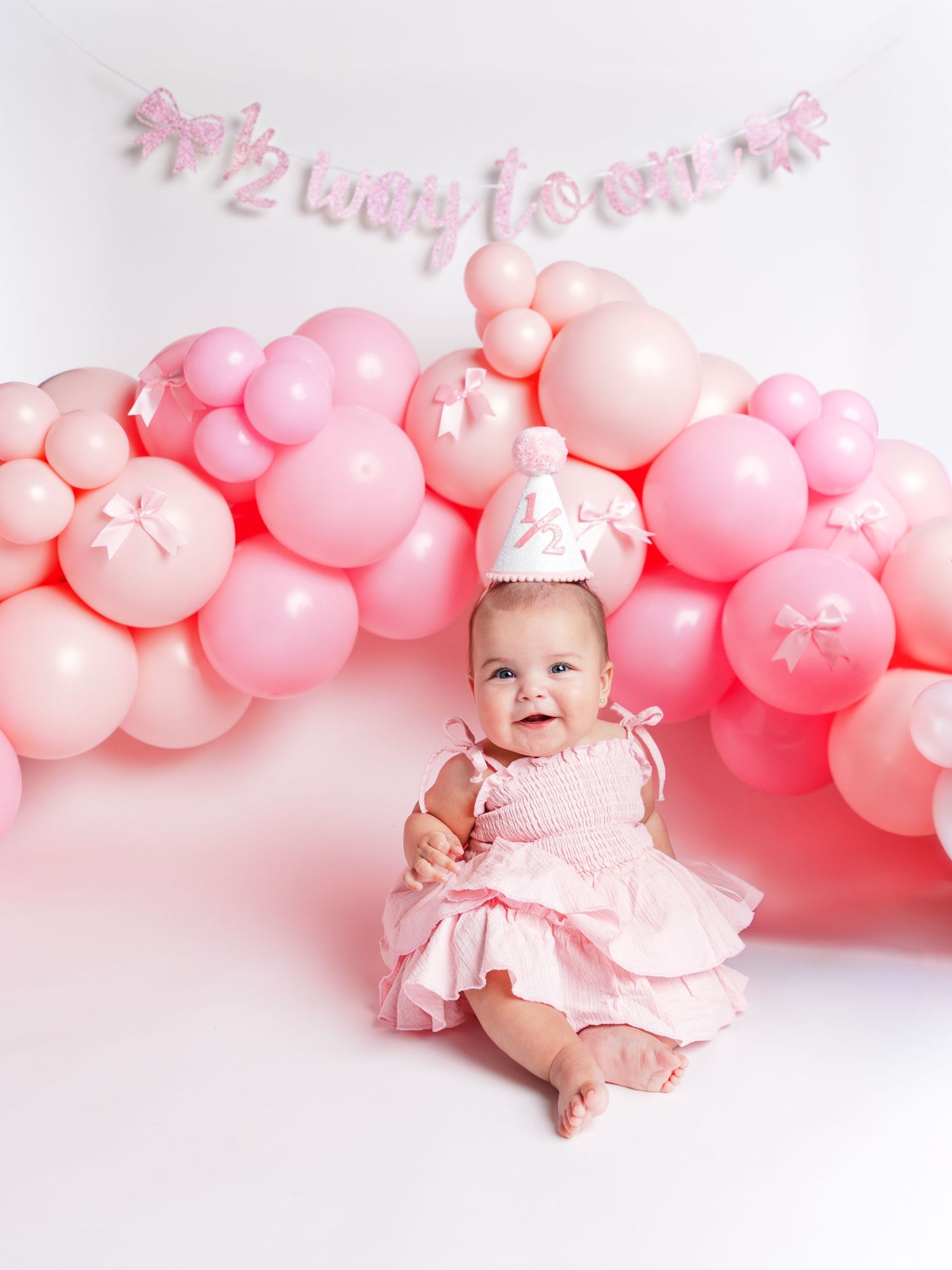 Baby girl in pink dress and crown sits before pink balloon arch celebrating her half birthday milestone.
