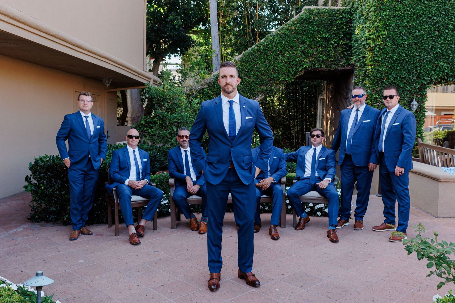 Groom stands confidently in navy blue suit with groomsmen posed behind him outdoors at wedding venue.