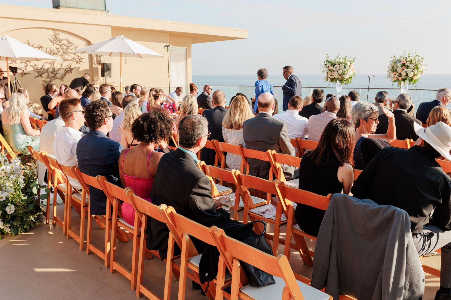 Outdoor wedding ceremony with guests seated on orange chairs overlooking the ocean on a sunny day.