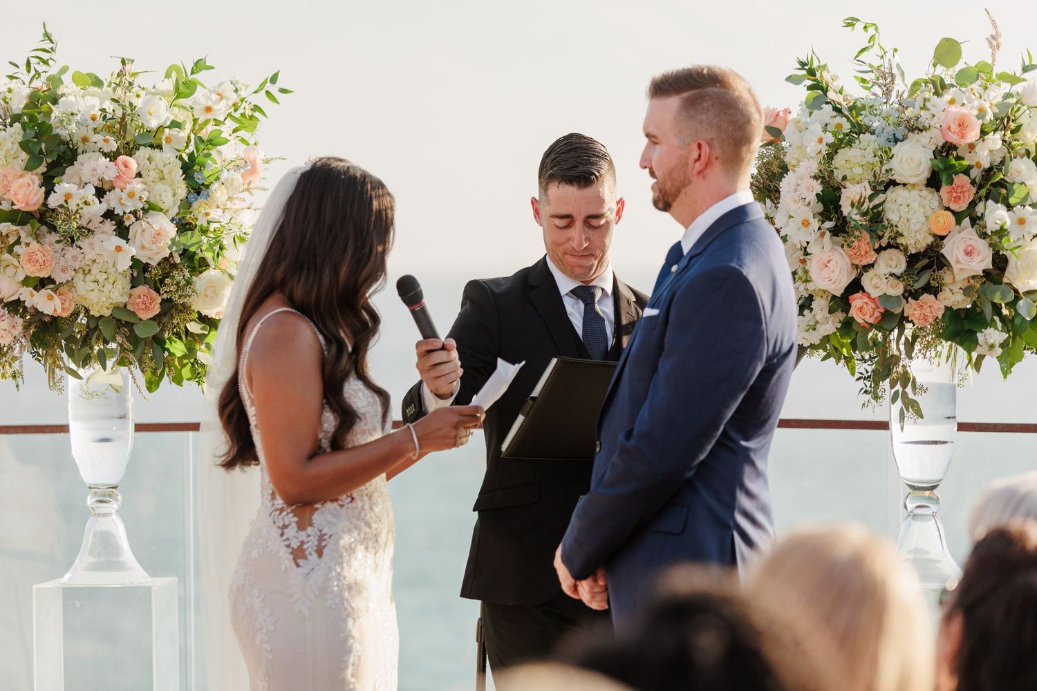 Officiant reading vows to smiling couple at elegant outdoor waterfront wedding with floral backdrop.