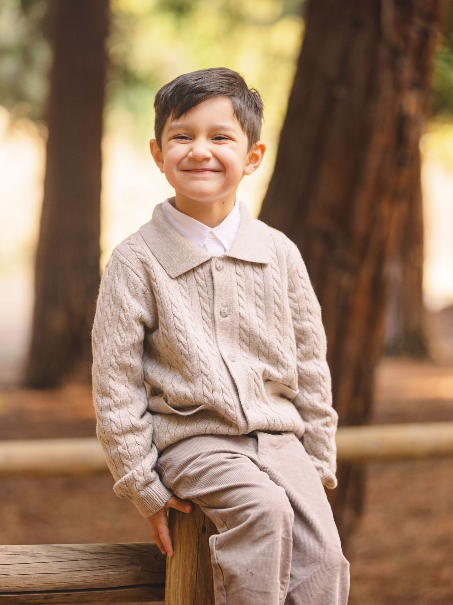 A young child in a beige sweater sits on a wooden bench in a forest setting with a bright smile.