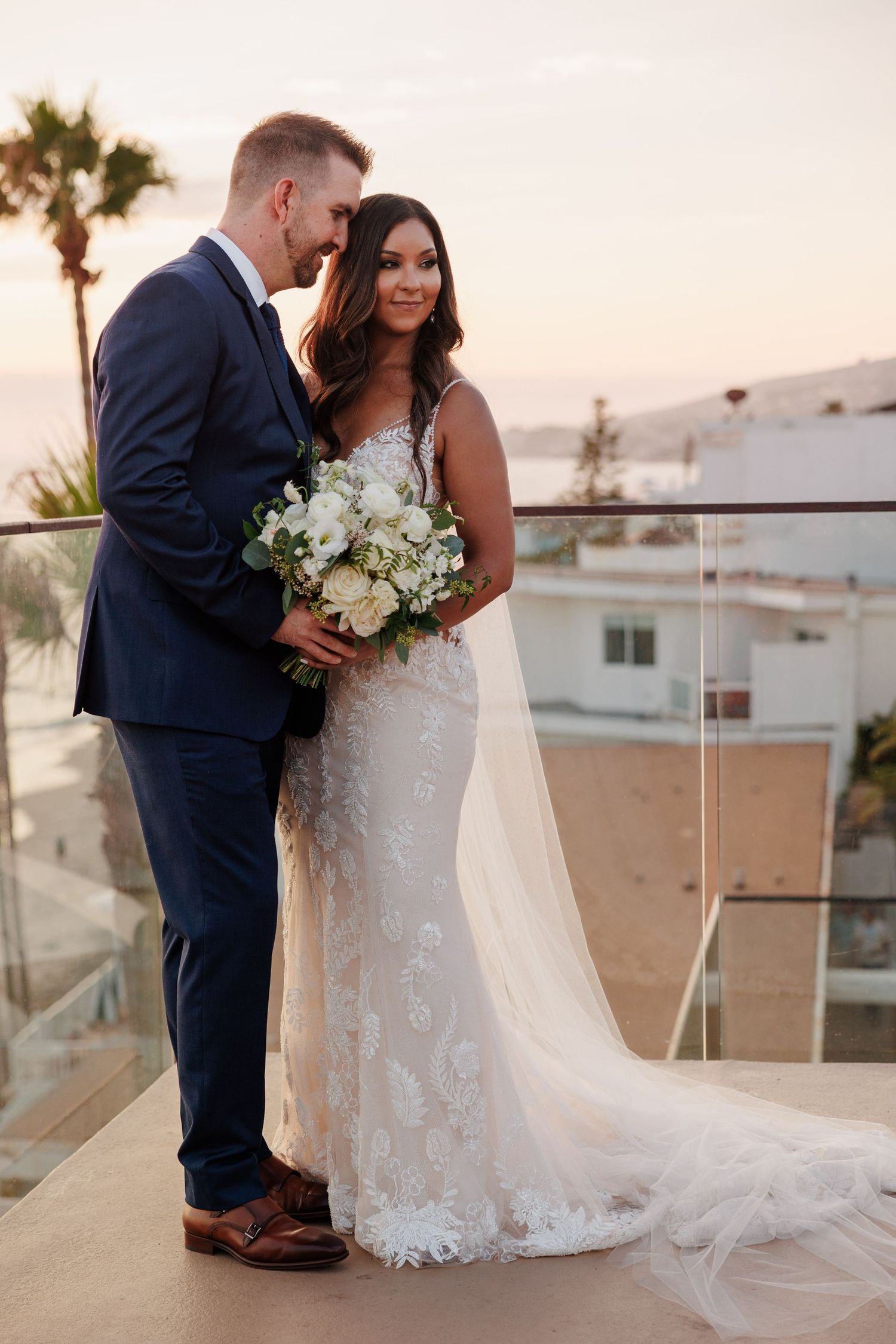 Bride in lace gown holds white bouquet with groom in navy suit on balcony at sunset.