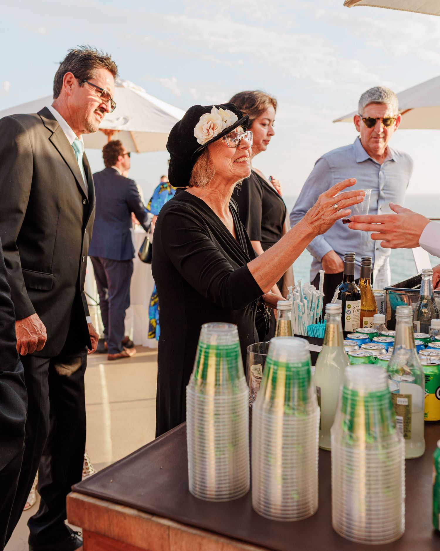 Guests gather at an outdoor bar with plastic cups and bottles during an elegant evening event under white tent canopies.