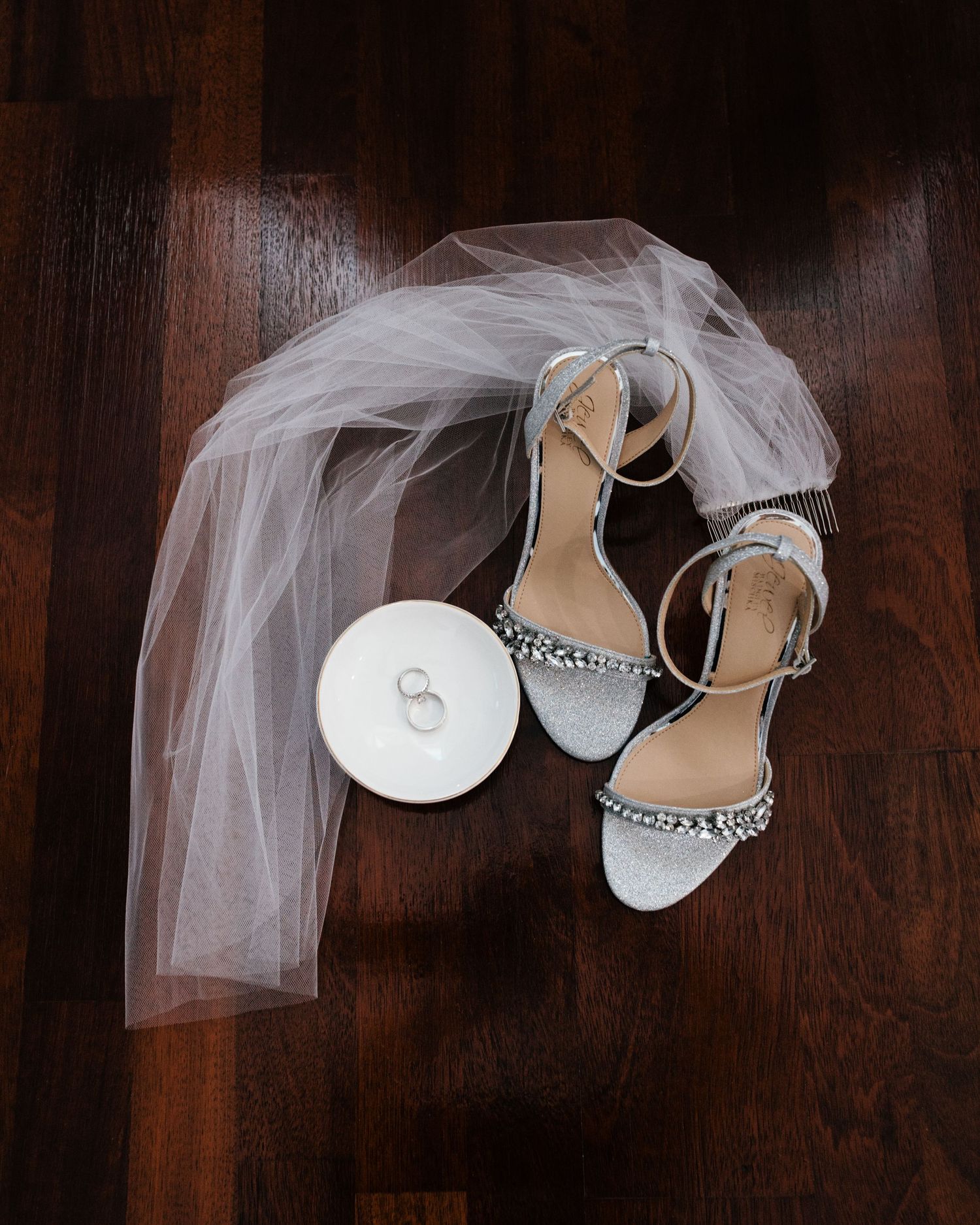 Silver glitter bridal heels with ankle straps beside a white ring dish with wedding bands and a flowing tulle veil on wood floor.
