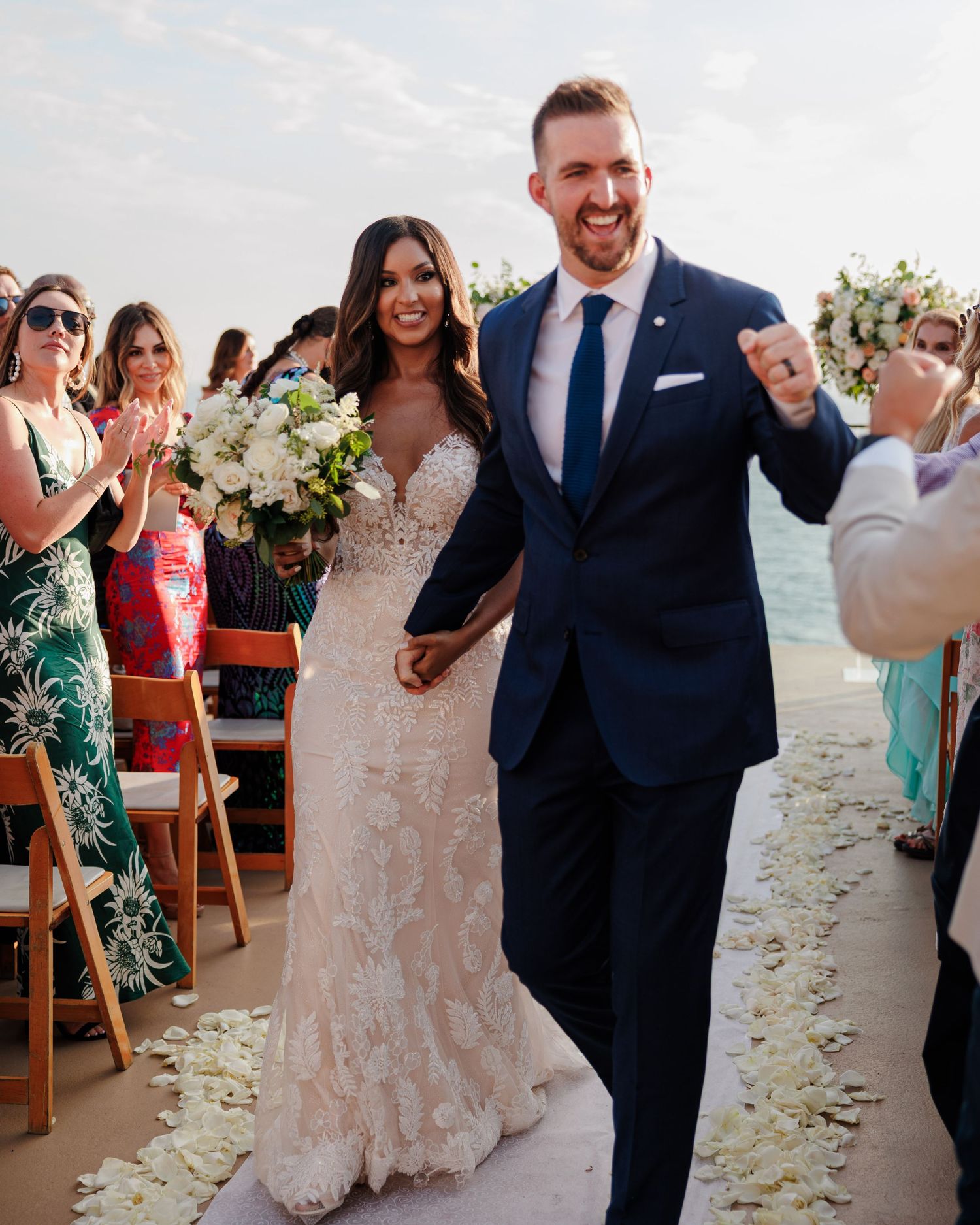Newlyweds walk down rose petal aisle as guests applaud at sunny outdoor beach wedding ceremony.