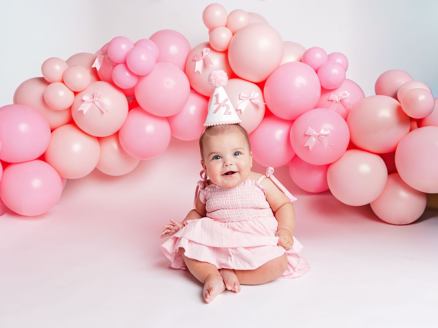 Baby girl in pink dress and birthday crown sits smiling before a pink balloon arch backdrop.