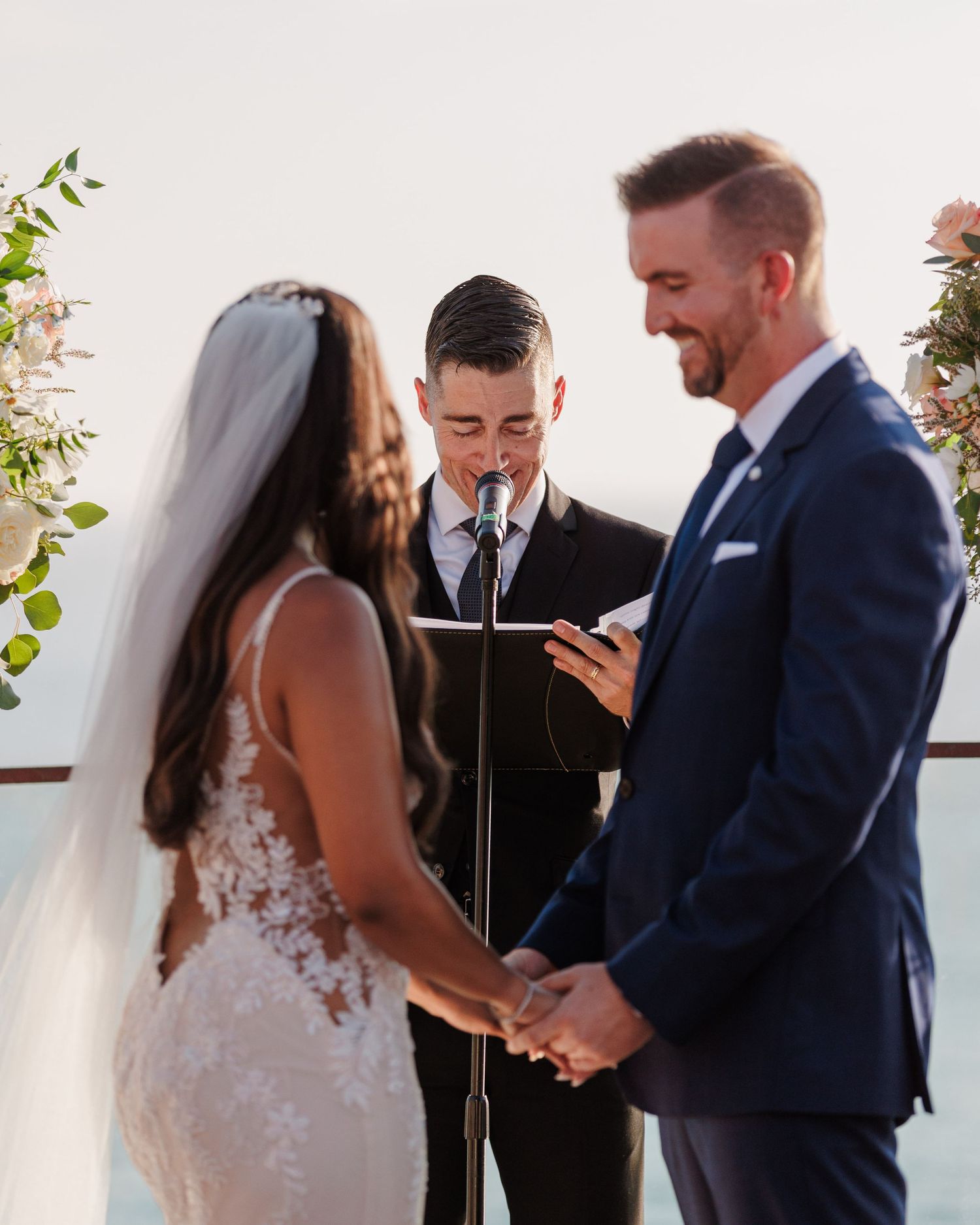 Bride and groom hold hands during outdoor wedding ceremony while officiant reads vows, floral arrangements in background.