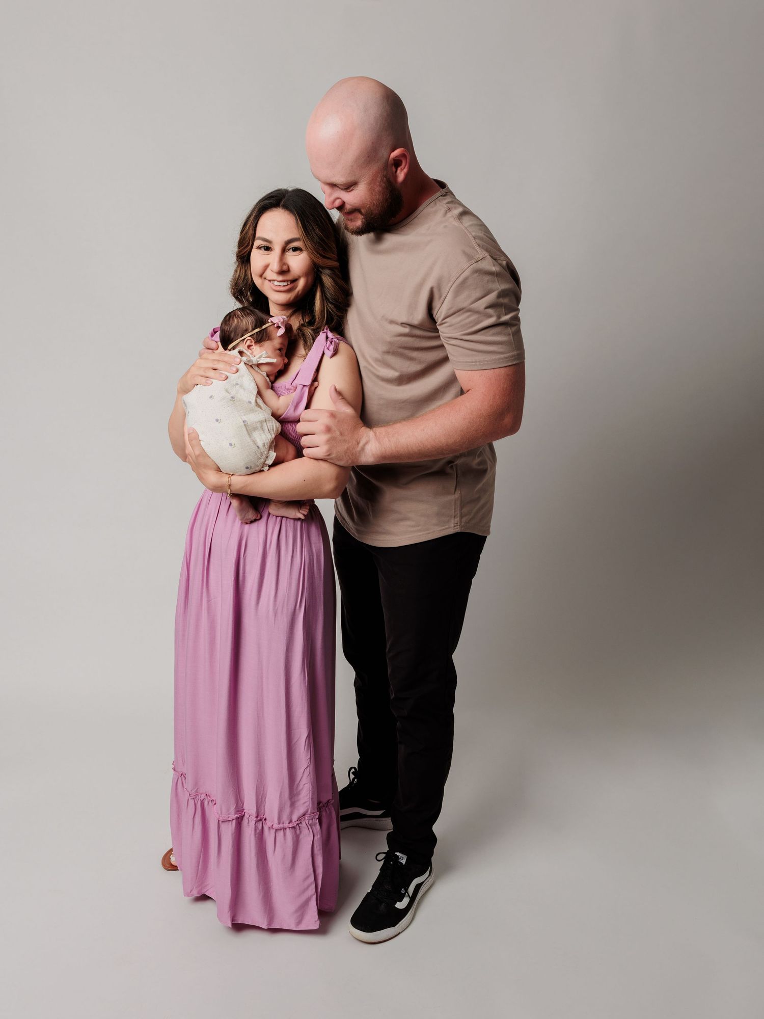 A couple in casual attire poses with their newborn baby against a light gray studio backdrop.