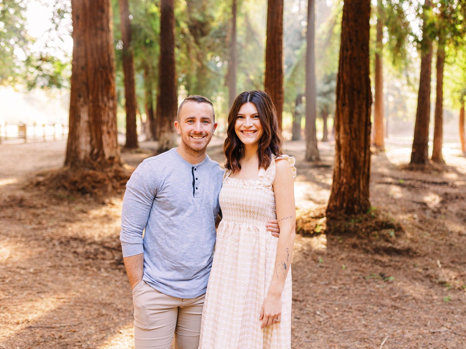 A couple stands together on a forest path with sunlight filtering through tall trees.