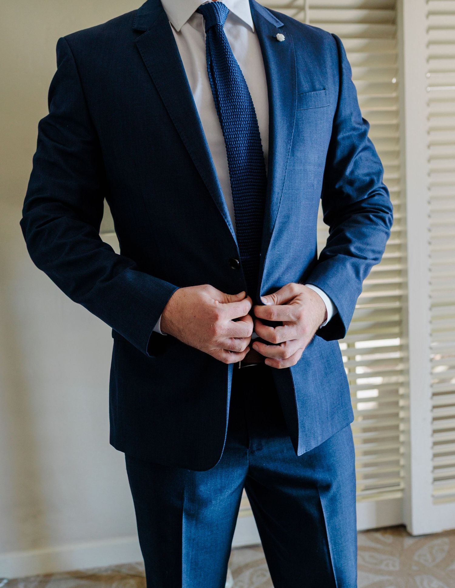 Groom in navy blue suit with blue tie buttoning jacket before wedding ceremony indoors.