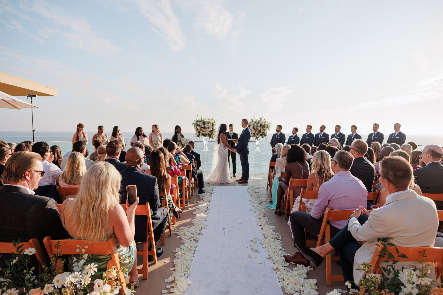 Outdoor beach wedding ceremony with guests seated on wooden chairs facing couple at floral altar with ocean backdrop.