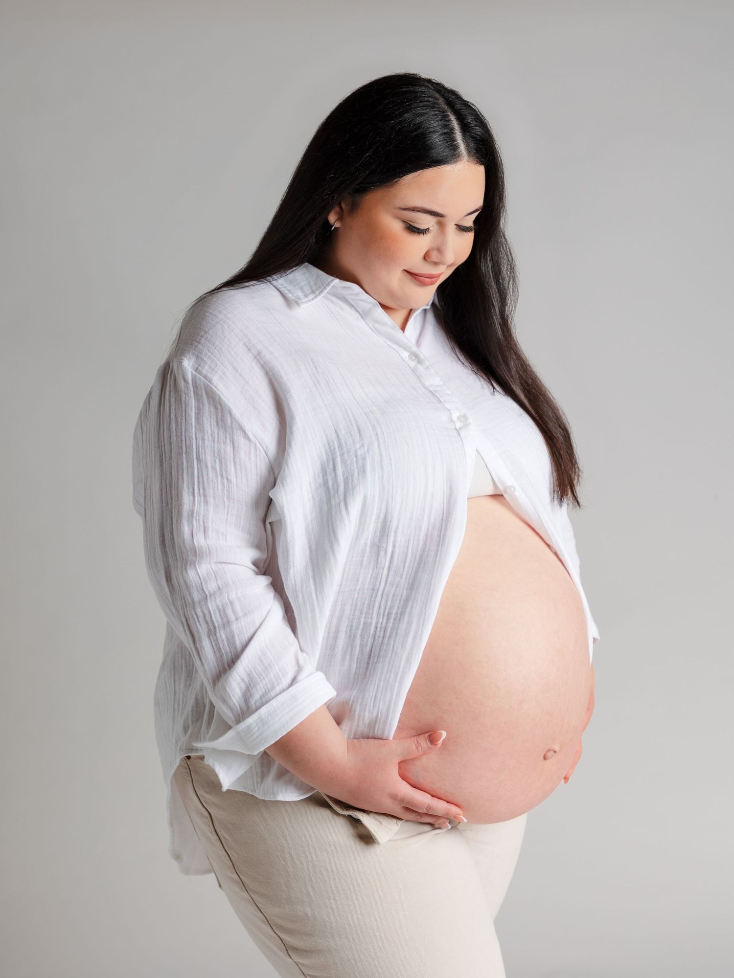 Pregnant woman in white linen shirt holding baby bump, maternity portrait on gray background.
