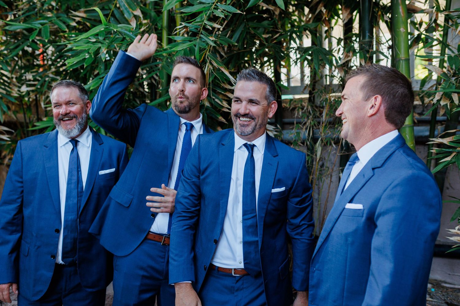 Four groomsmen in blue suits laughing outdoors near bamboo.