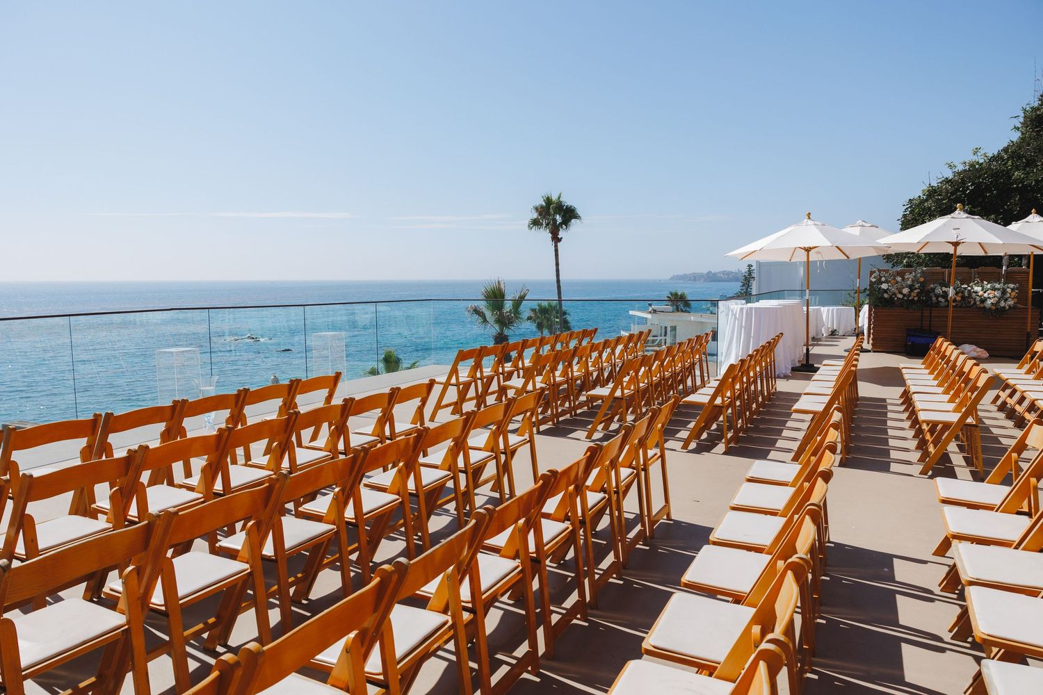 Outdoor wedding ceremony setup with wooden chairs facing ocean view, palm tree, and white umbrellas under clear blue sky.