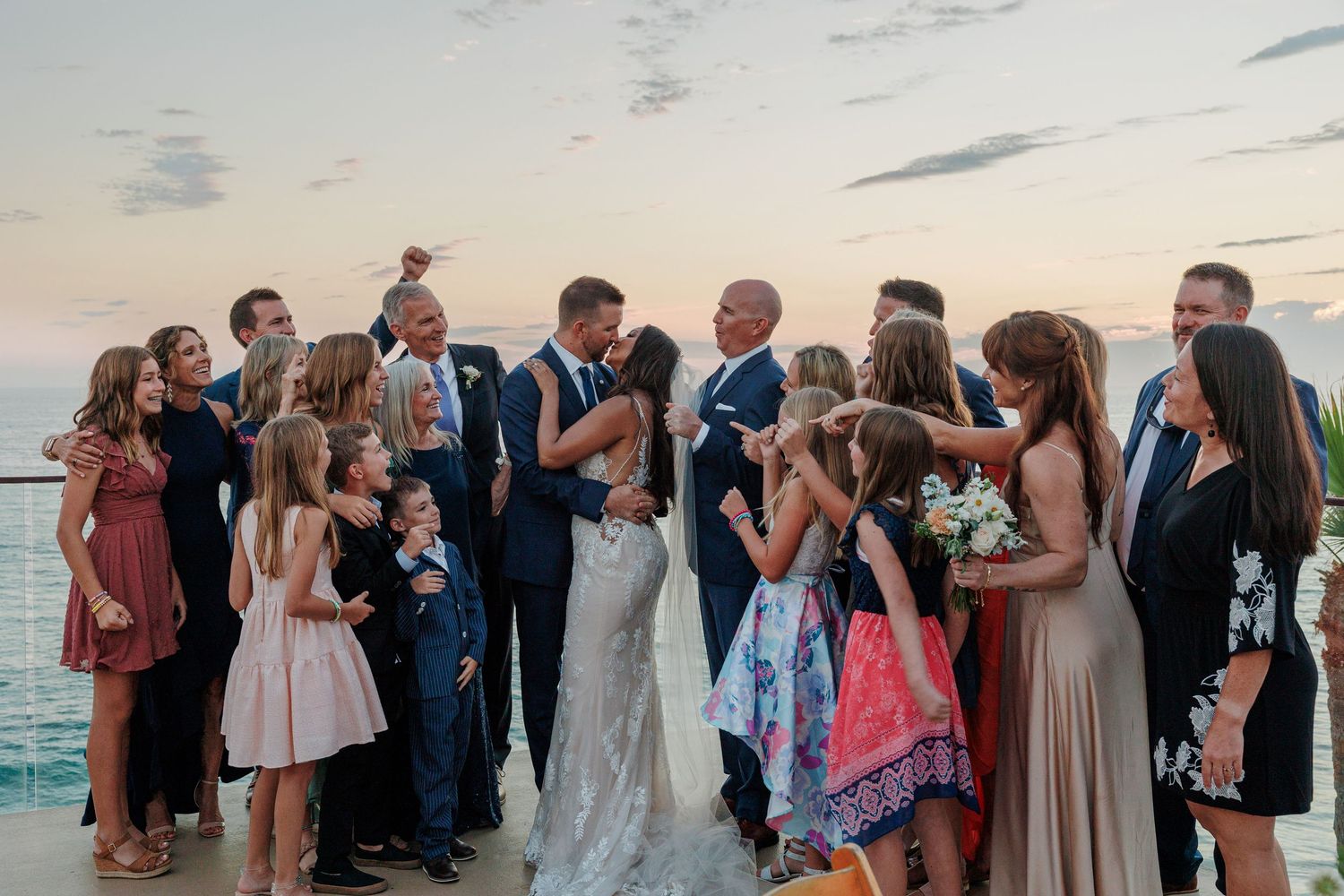 Bride and groom share a kiss surrounded by cheering wedding guests at oceanside reception.