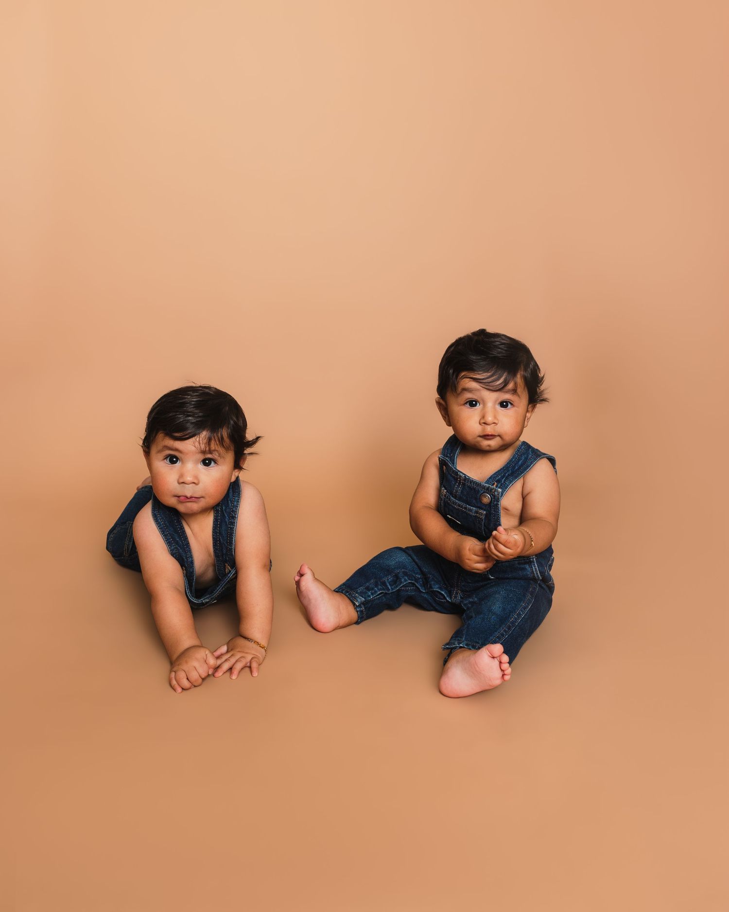 Twin toddlers in matching blue denim overalls pose on peachy-brown studio backdrop.
