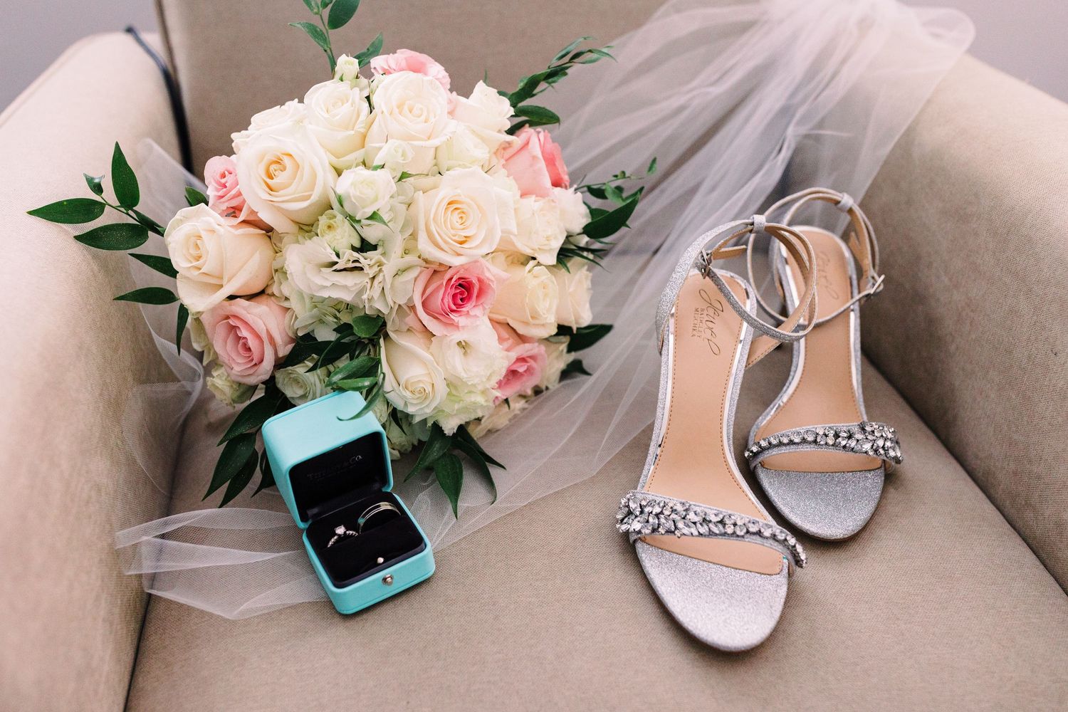Bridal details flat lay featuring a pink and white rose bouquet, silver heeled sandals, and a teal ring box on a beige chair.