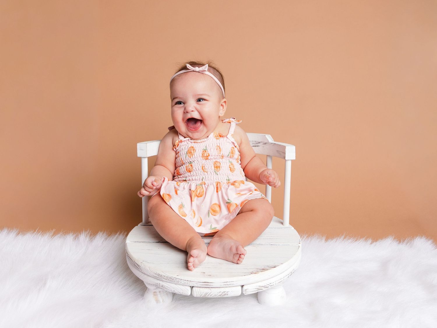 Laughing baby girl in floral dress sits on white chair against tan background.