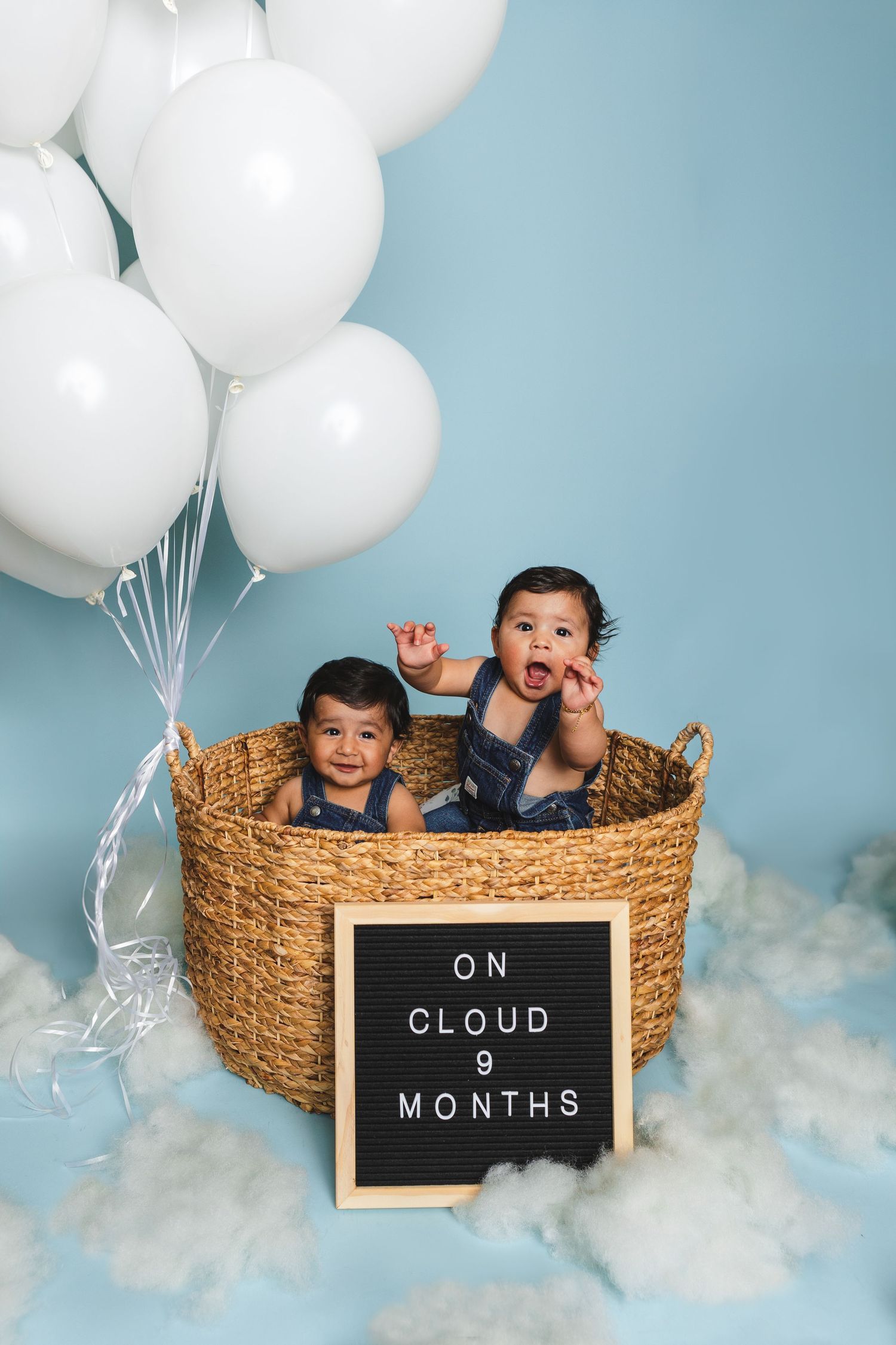 A whimsical 9-month baby photo setup with white balloons, clouds, and a woven basket against a blue backdrop.