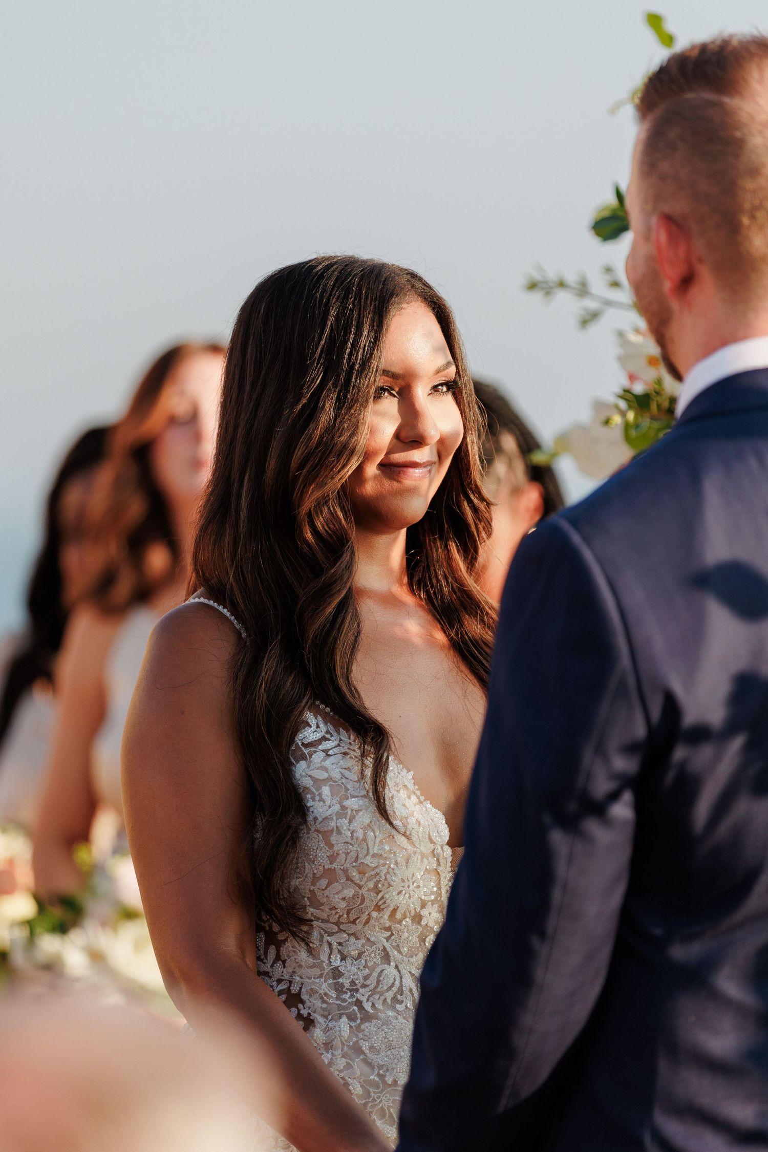 Beautiful bride with long dark hair in lace gown smiling at groom during outdoor wedding ceremony.