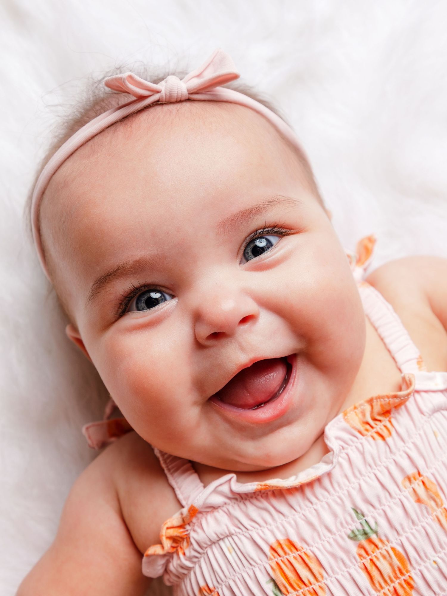 Smiling baby girl wearing a pink smocked dress and bow headband, lying on a white fluffy blanket.