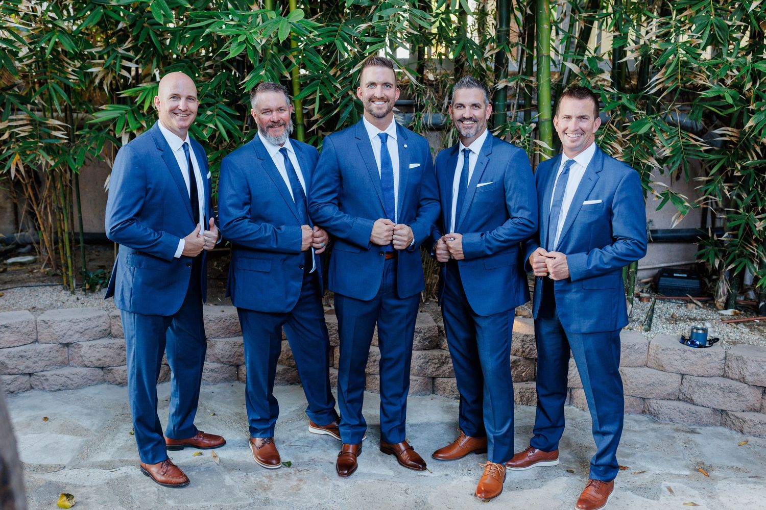 Five groomsmen in matching blue suits and brown shoes pose together outdoors at a wedding.
