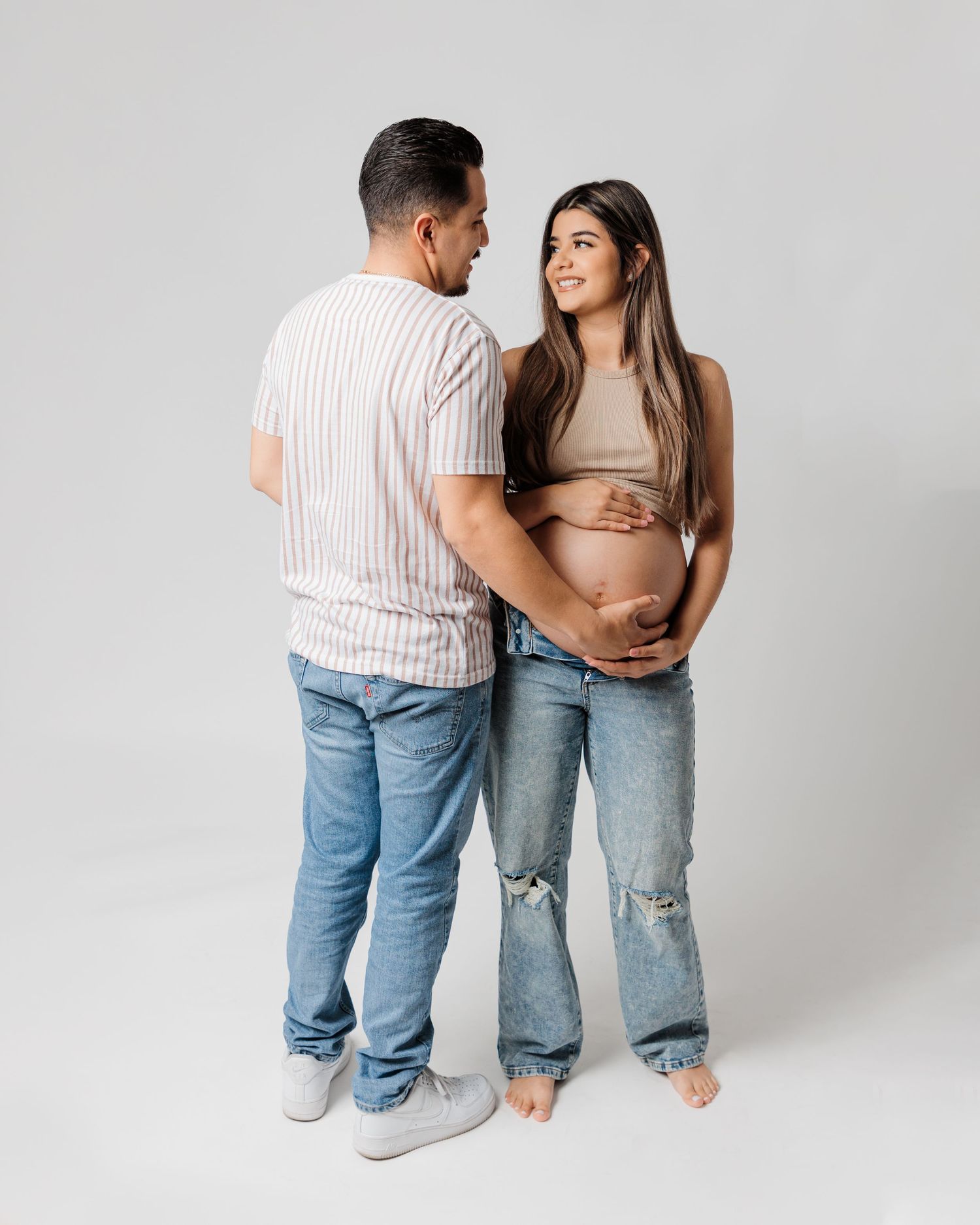 Standing couple shares loving moment during maternity photoshoot on white background.