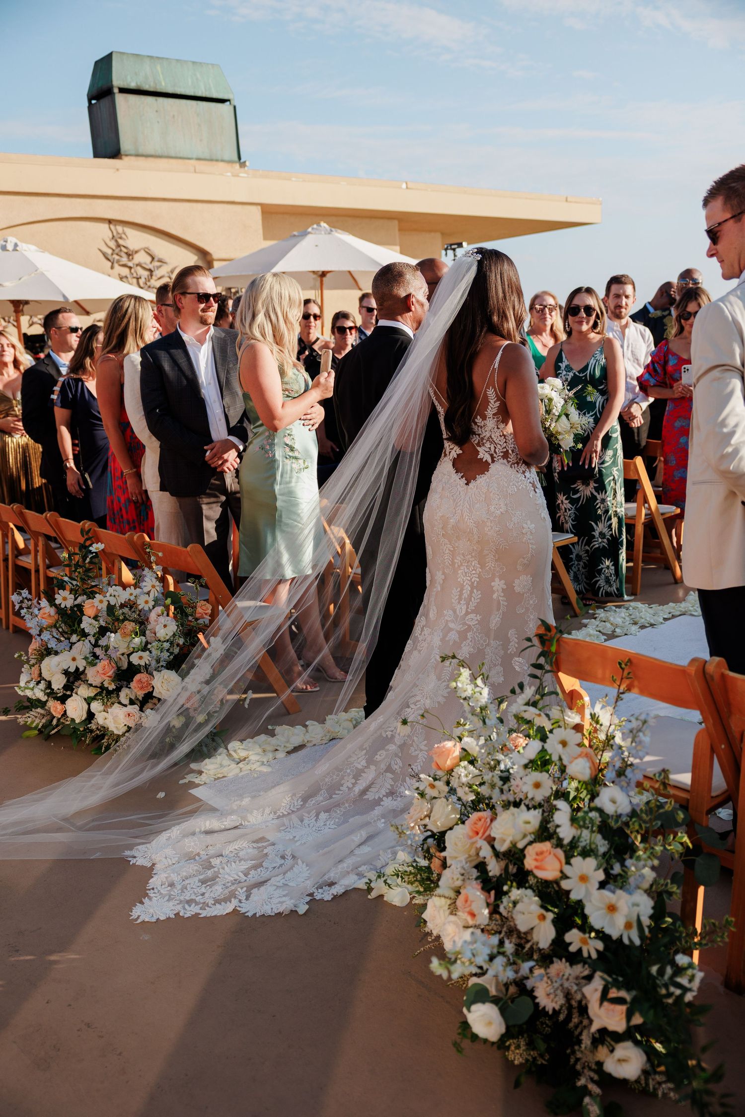 Bride in lace gown with cathedral veil stands at outdoor wedding altar surrounded by floral arrangements.