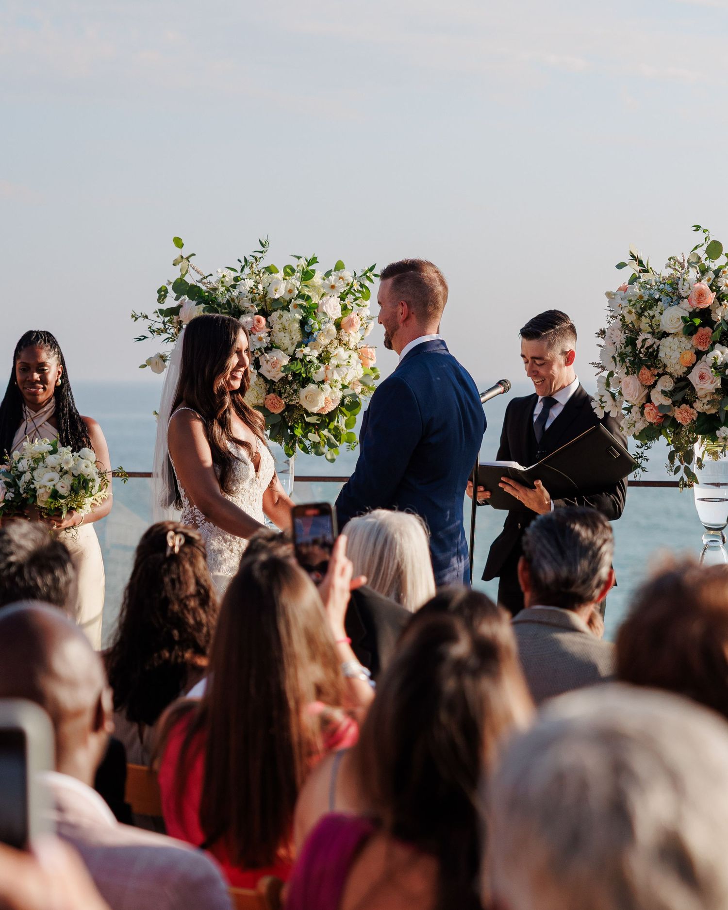Bride and groom exchange vows at oceanfront altar adorned with pink and white floral arrangements.