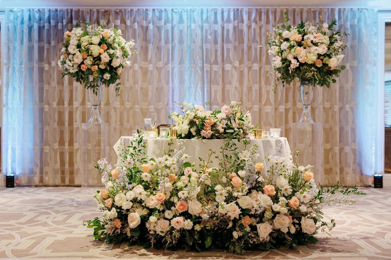 Elegant wedding sweetheart table adorned with lush white and peach floral arrangements against a shimmering draped backdrop.