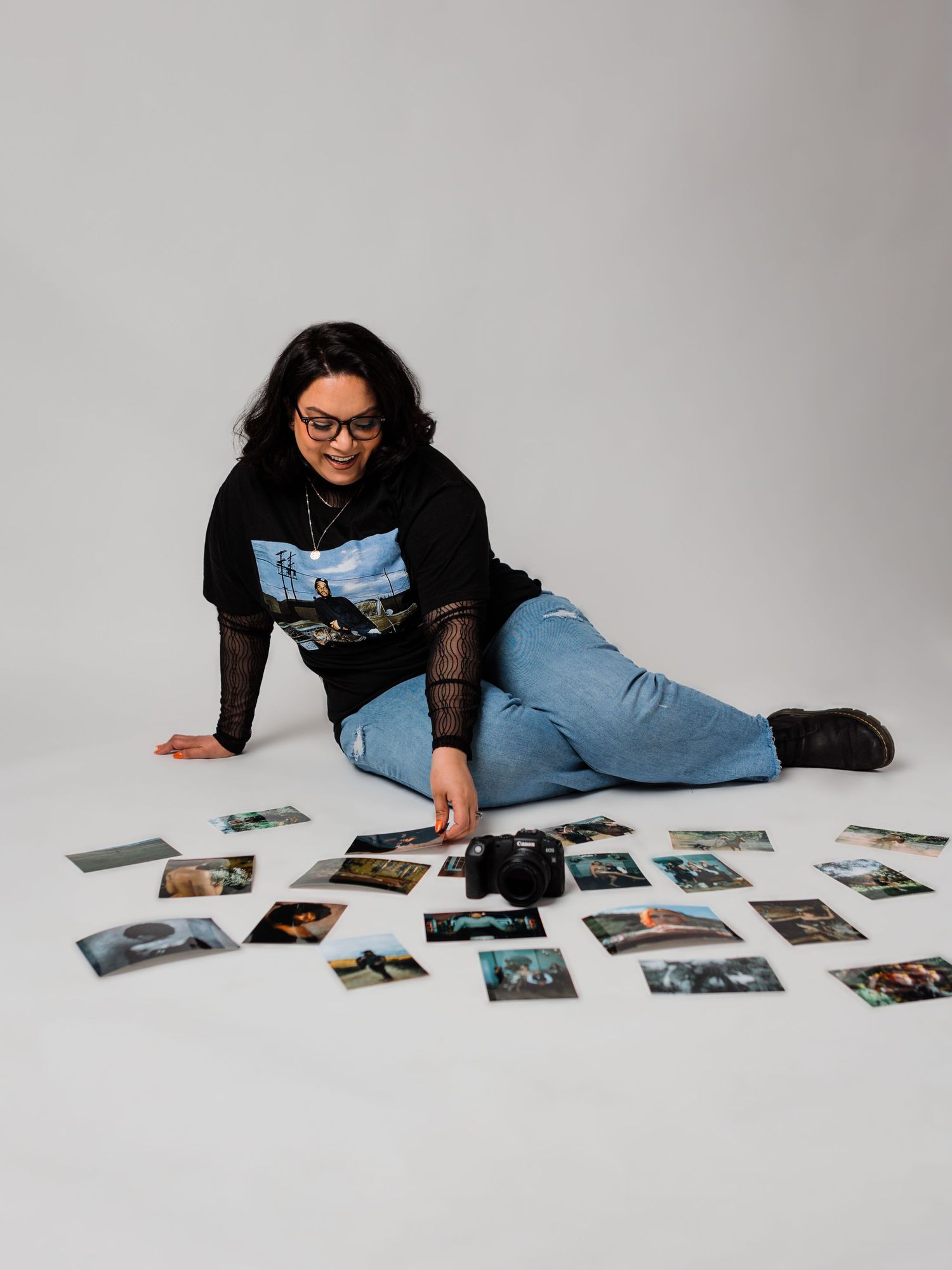Woman sitting on floor surrounded by scattered photos, wearing black graphic tee and jeans, reaching toward a camera.