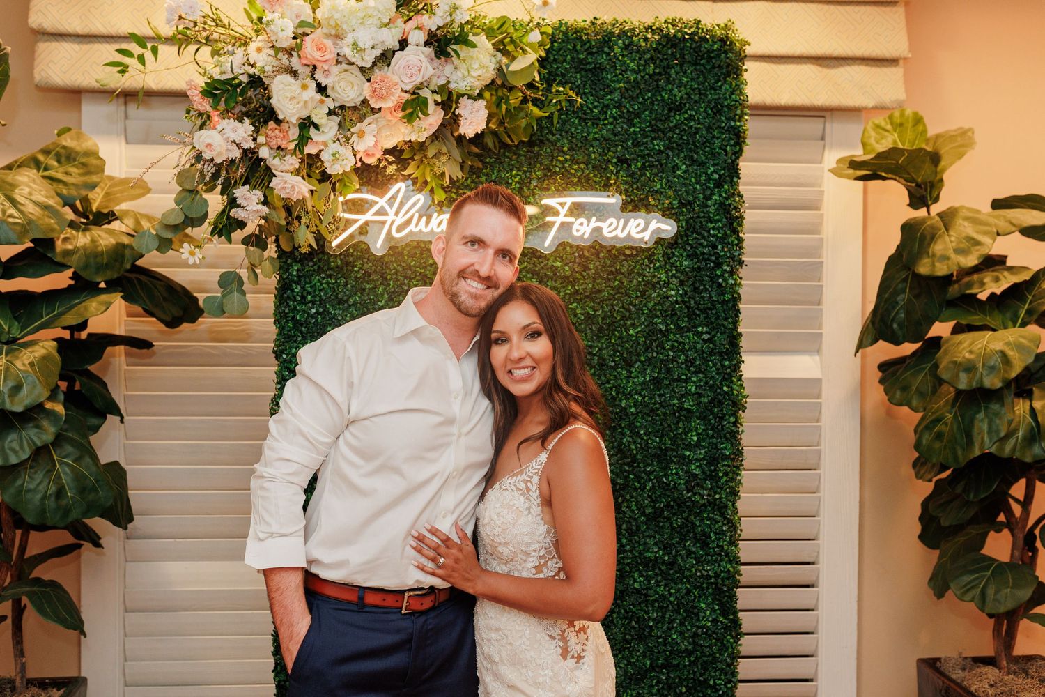 Bride and groom pose together in front of an Always and Forever neon sign and floral wall.