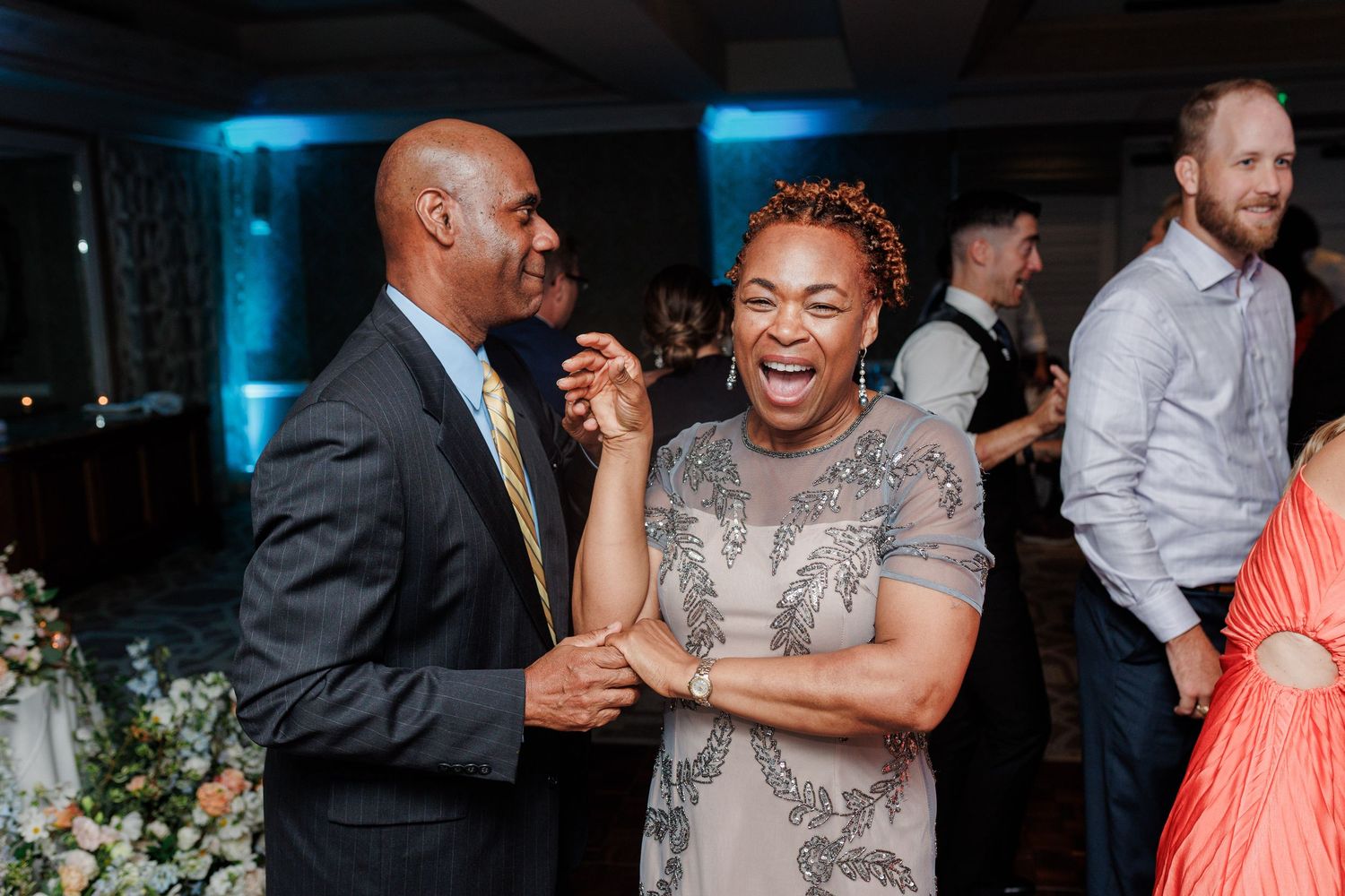 Joyful couple dancing and laughing at a wedding reception with guests in the background.