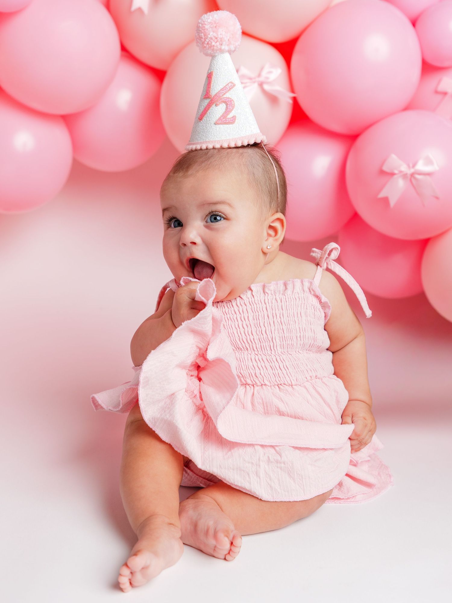 A baby girl in a pink smocked romper and birthday party hat sits in front of a pink balloon arch.