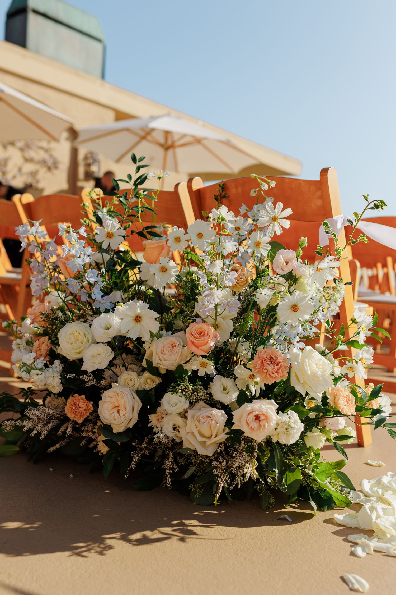Elegant outdoor wedding floral arrangement with white daisies, peach roses, and greenery on sandy ground.