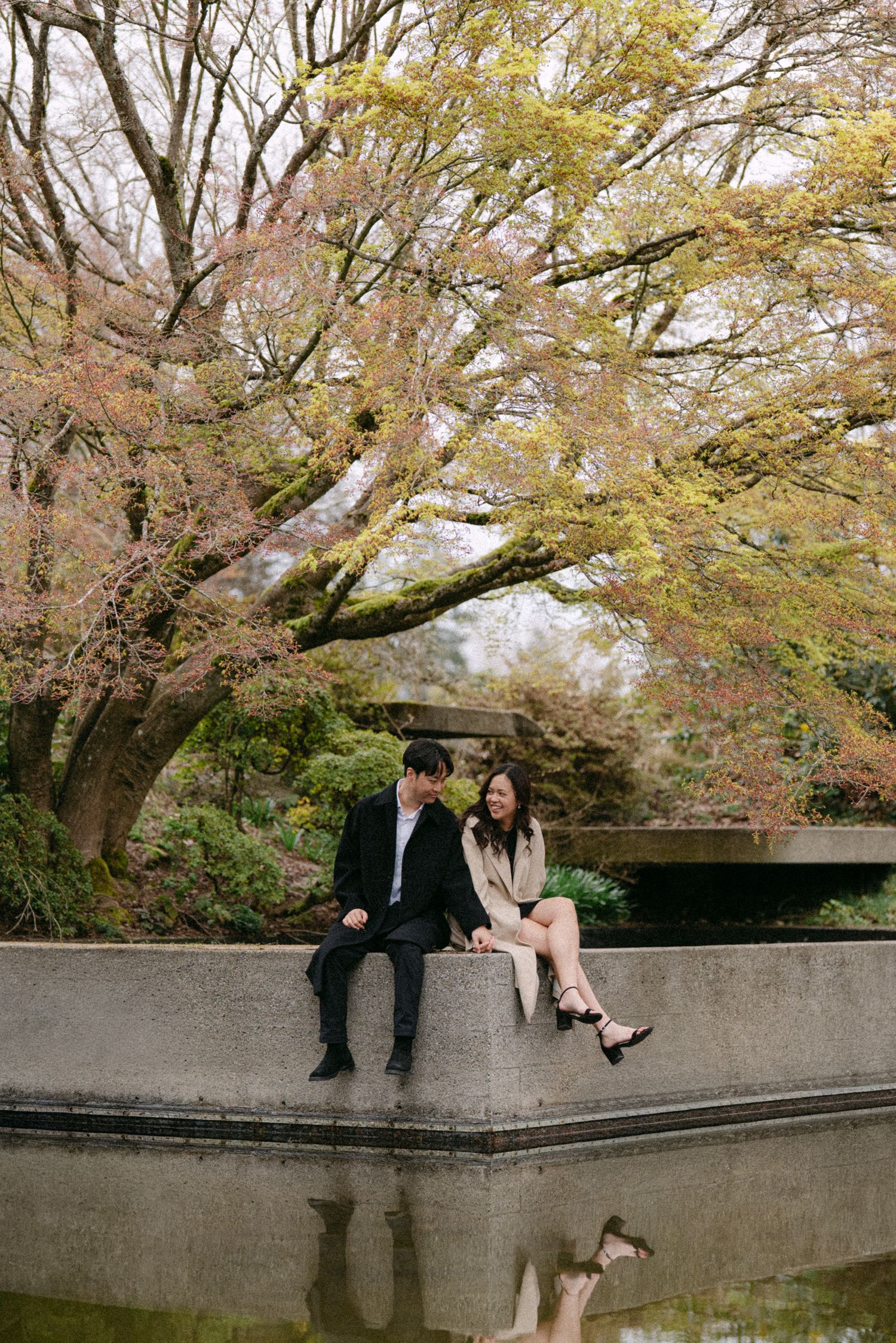 Couple sitting and laughing in front of pond at UBC Rose Garden. Vancouver engagement by Victor Li Photography.