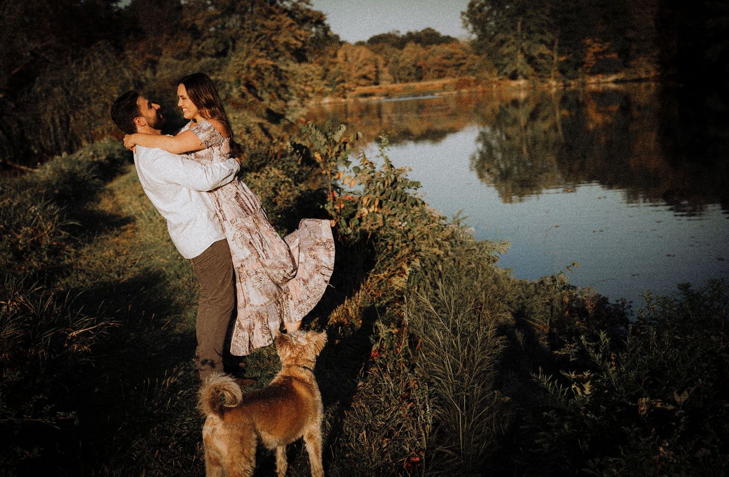 A romantic couple embraces by a serene lake at sunset while their dog watches from below.