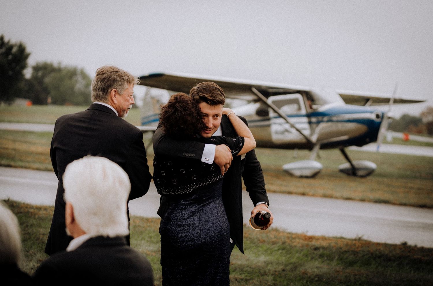 Wedding party poses by a small aircraft on a grassy airfield.