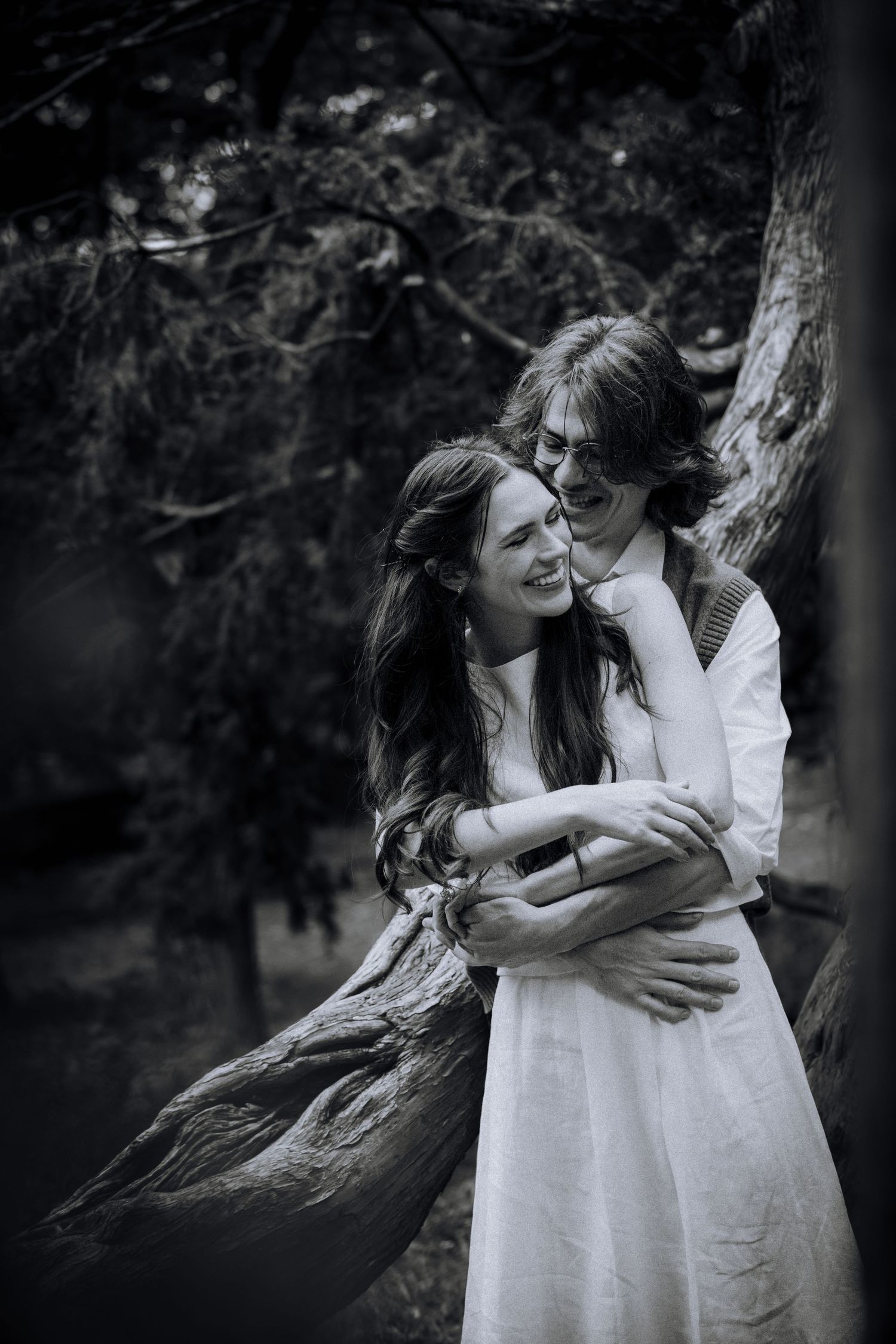 A couple embraces and laughs together while standing by a twisted tree trunk in a moody black and white setting.