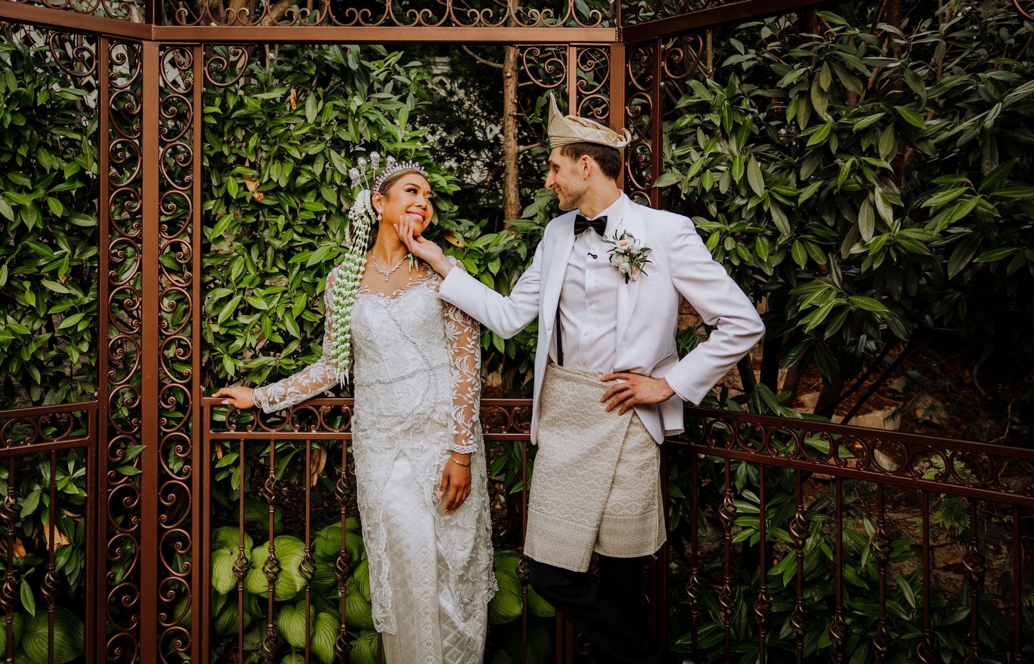 A couple in elegant white wedding attire pose together under a rustic garden arbor with lush greenery.