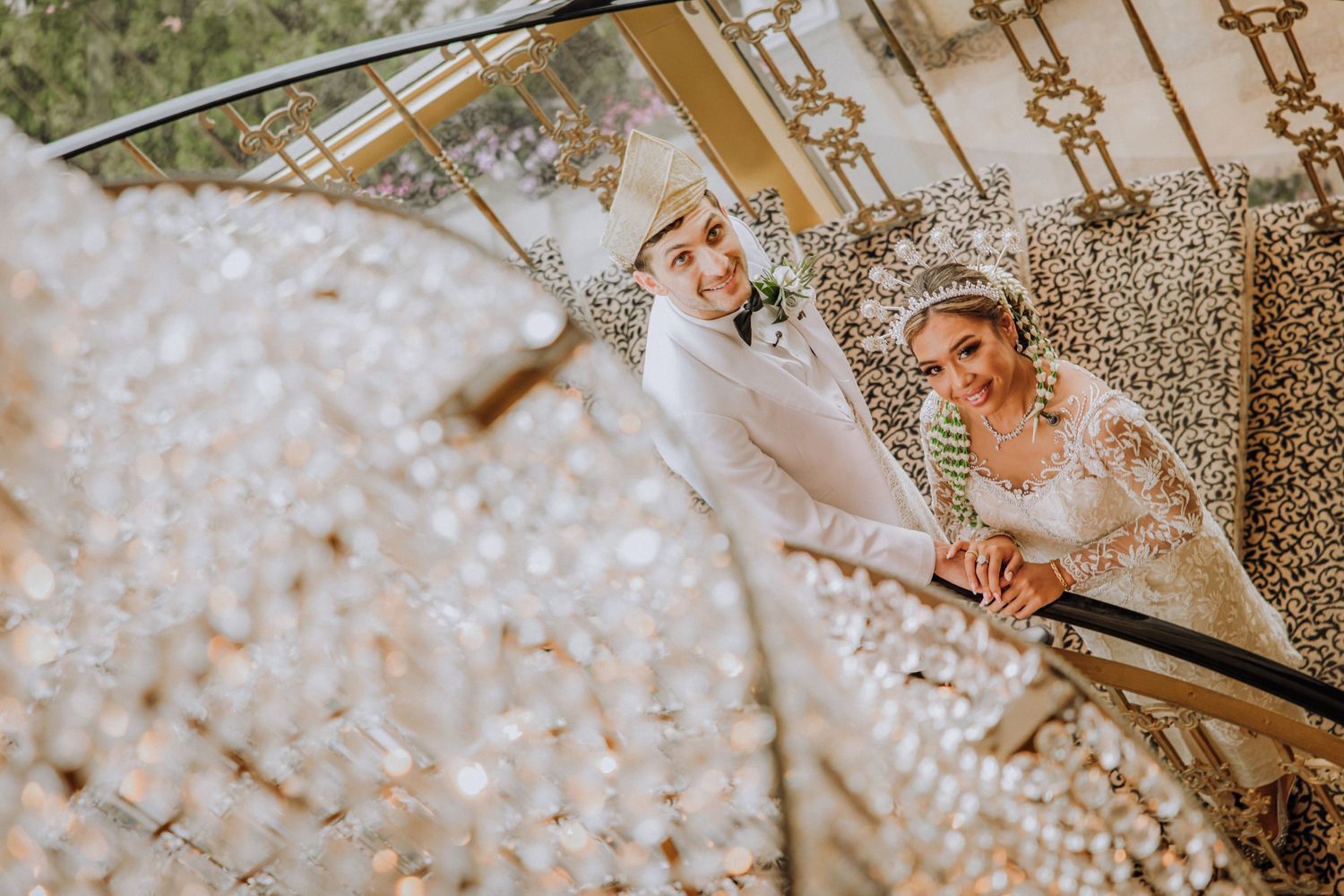 Two people in elegant white attire smile toward each other in a glamorous setting with crystal decorations and warm lighting.