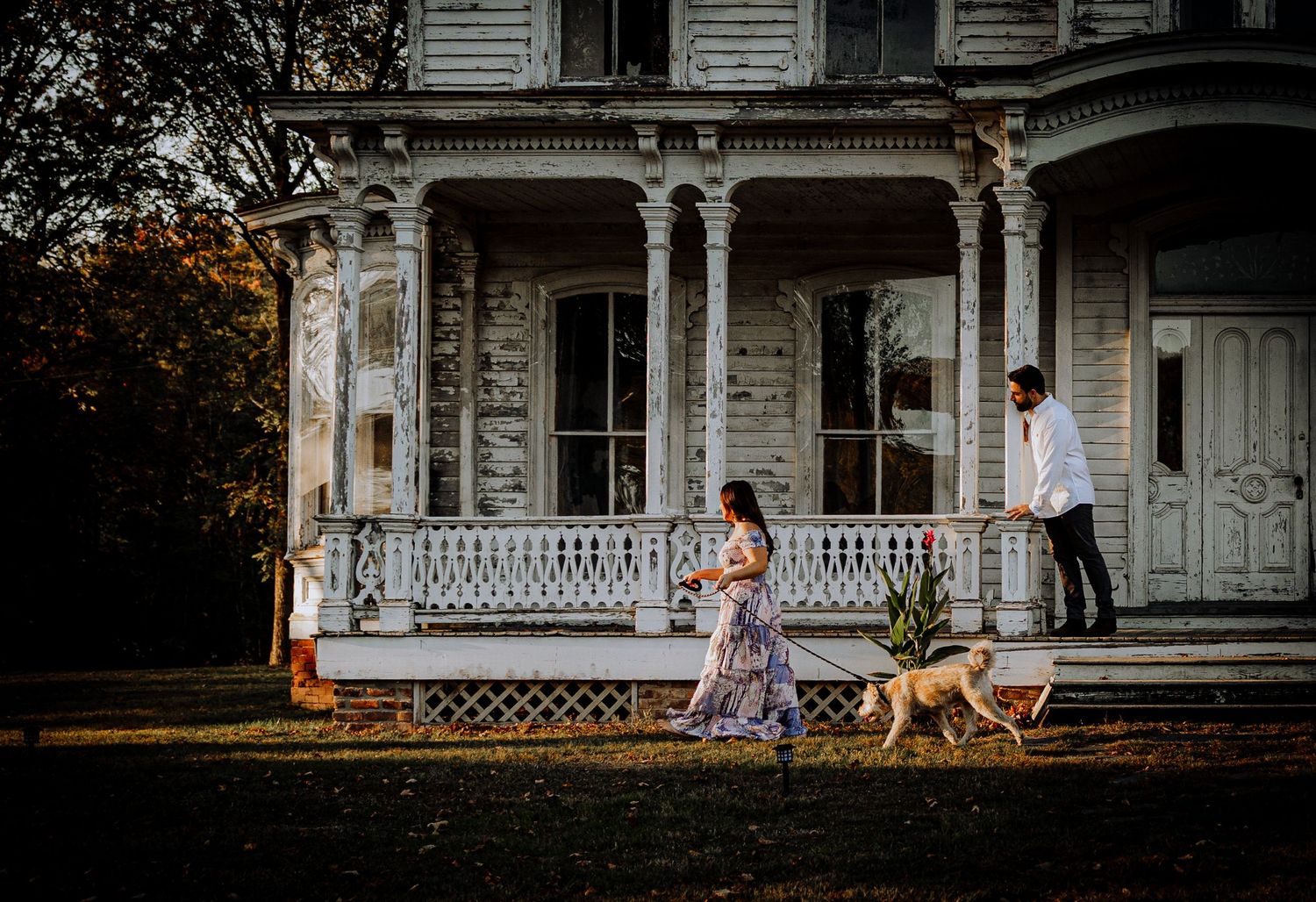 A couple and their dog walk past a vintage Victorian house with ornate white trim.