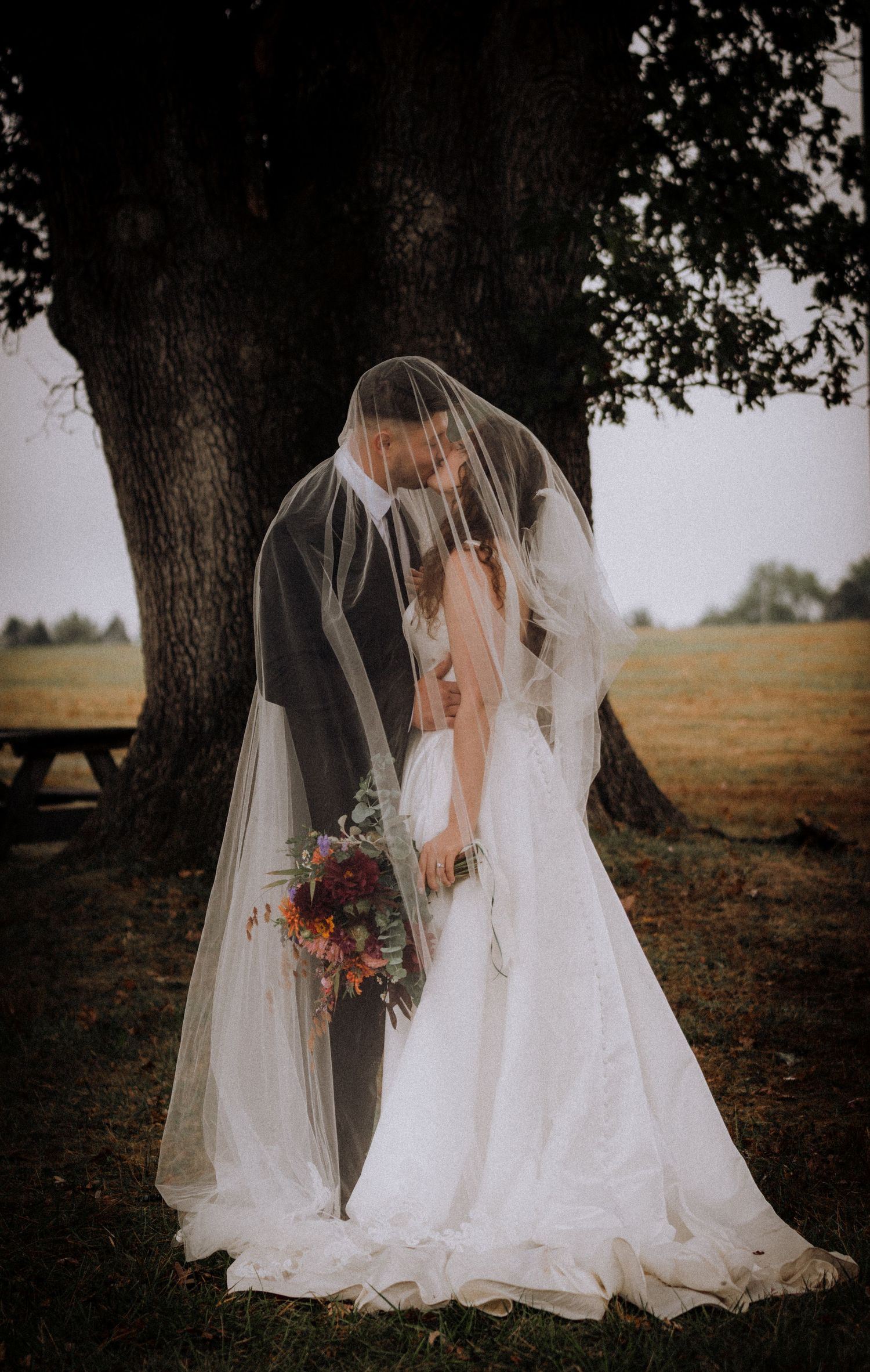 Two wedding dresses with veils photographed under trees in an autumn field.