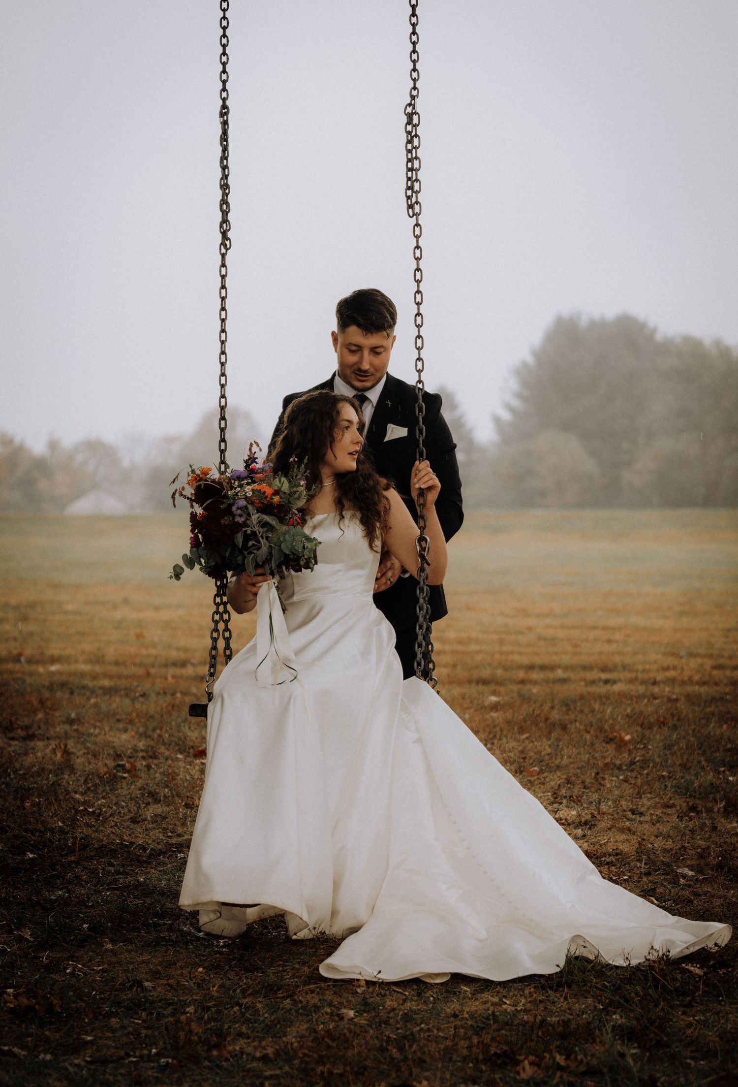 A couple shares romantic moments on a swing in a misty field during their wedding photoshoot.