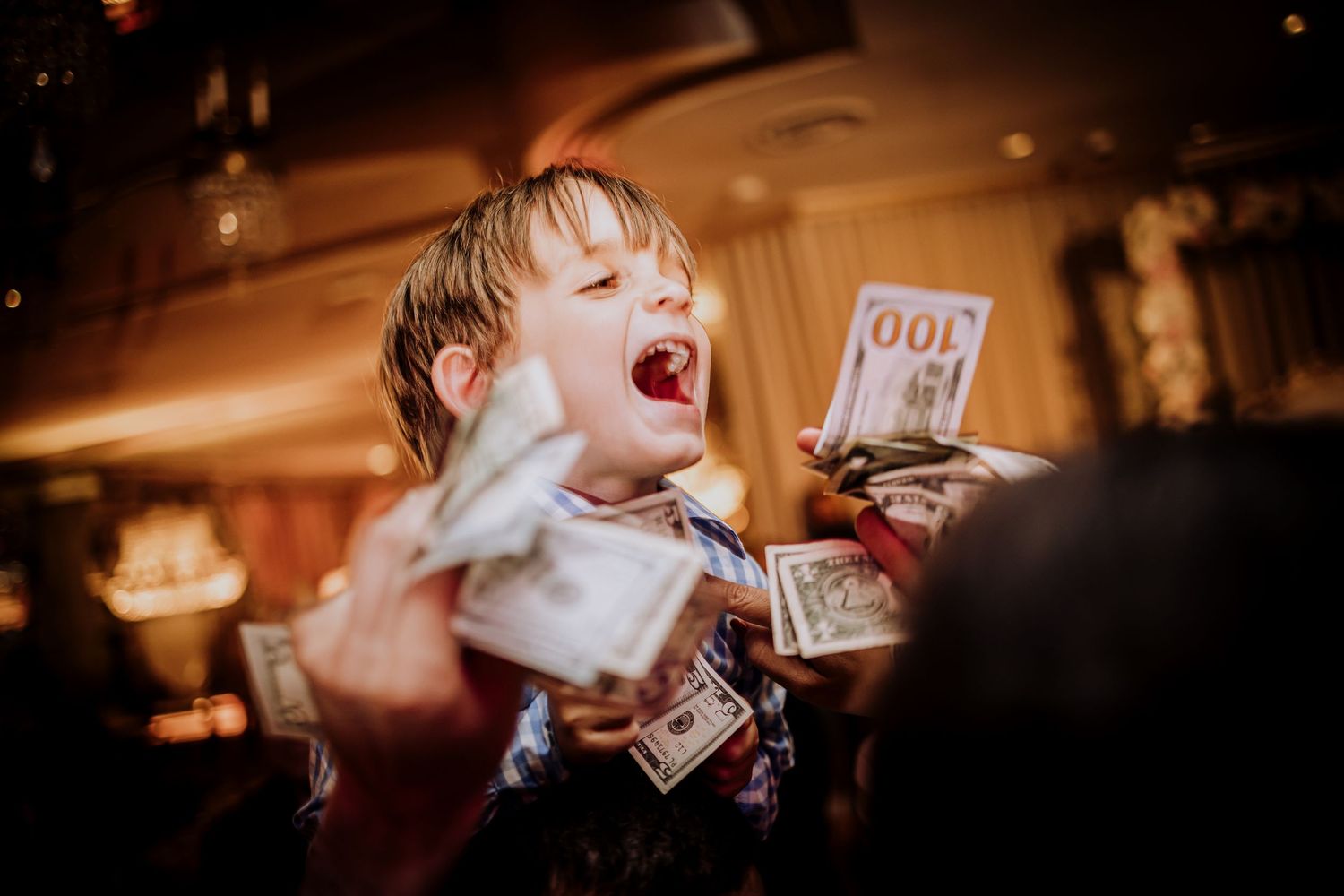 Young child in white laughs excitedly while holding money in a warmly lit room with a blurred background.