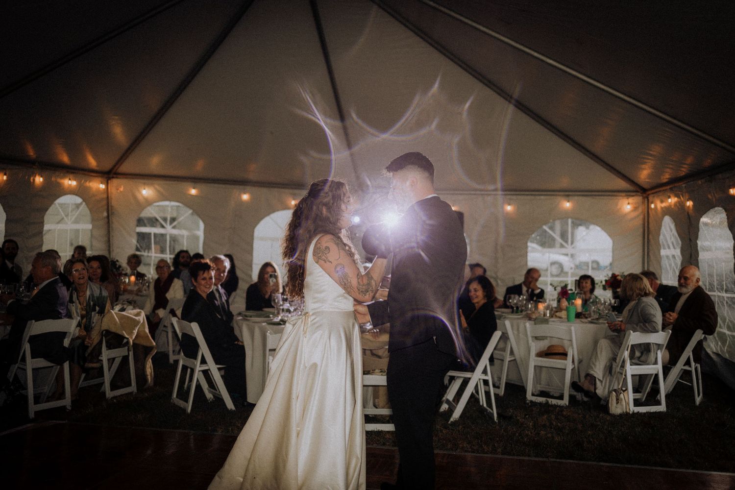 Wedding reception tent illuminated at night with guests seated at round tables.
