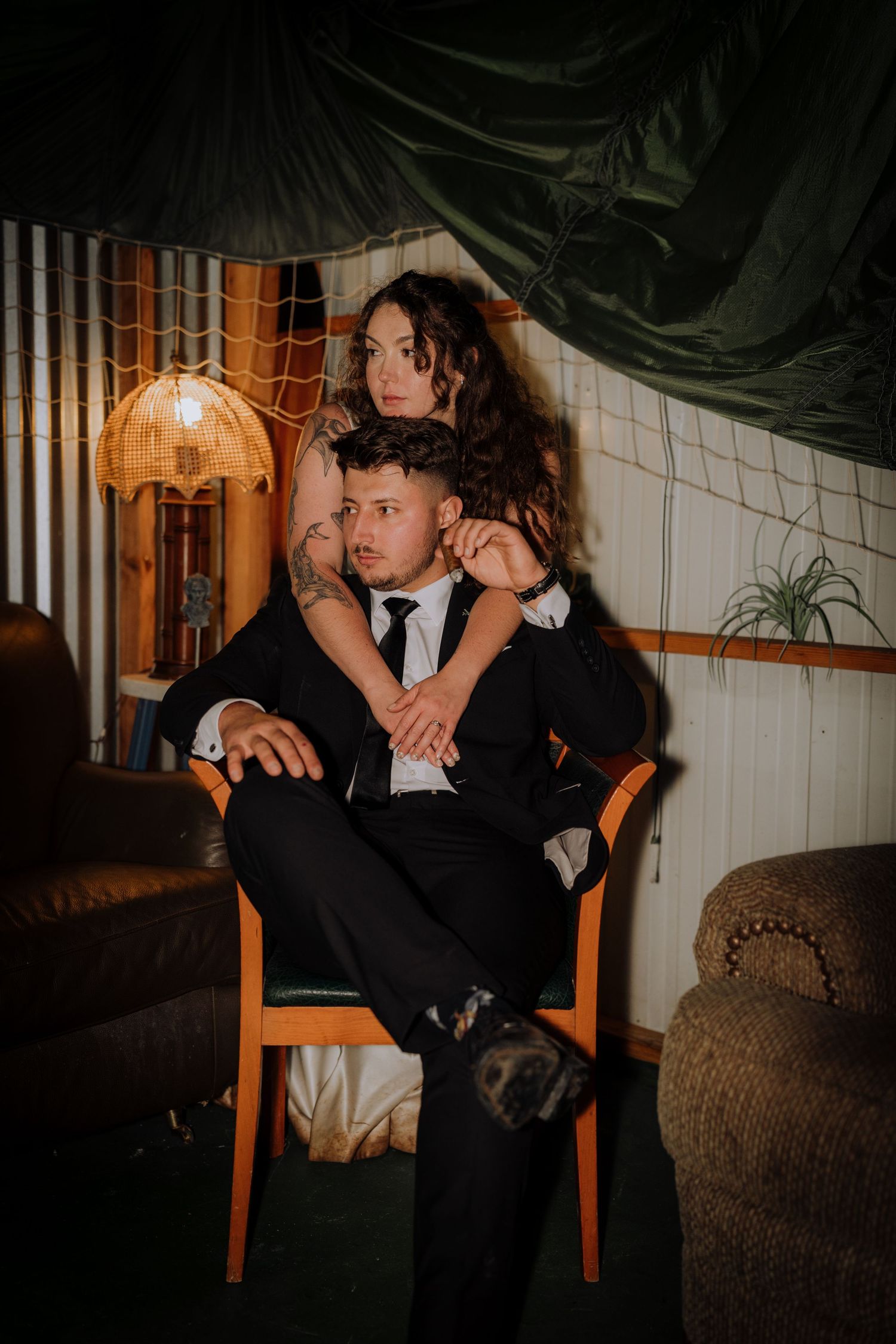A couple in formal attire share an intimate moment in a dimly lit room with wooden chairs and dark decor.