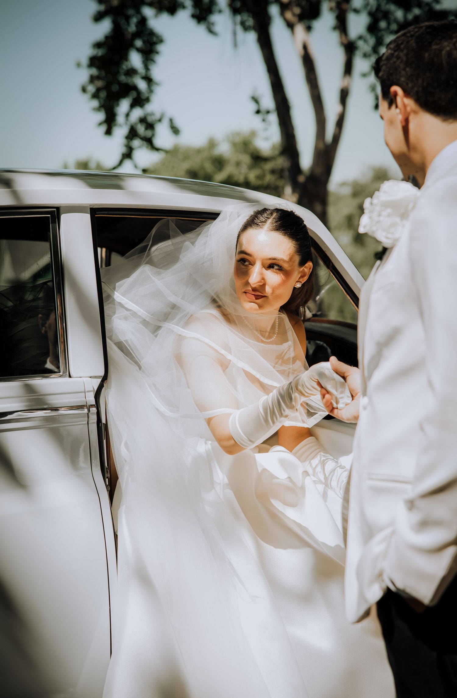 A classic white wedding car scene showing the bride's flowing dress and veil as she enters the vehicle.
